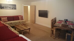 Ein Wohnzimmer mit einer Couch, einem Couchtisch, einem Esstisch mit Stühlen, einem an der Wand befestigten Fernseher und einer Tür, die in einen anderen Raum führt.