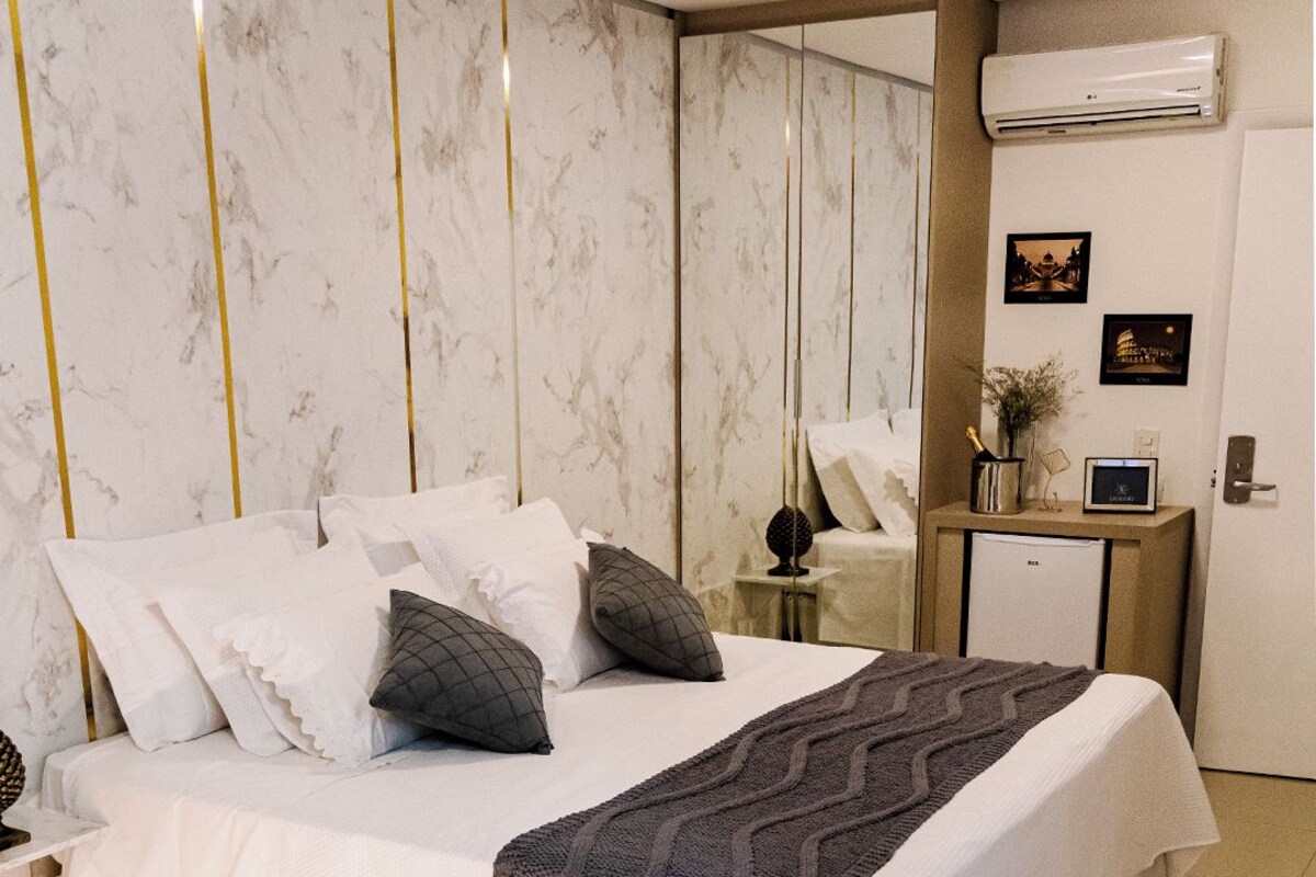 Una habitación de hotel moderna con una cama grande, una mesita de noche con lámpara, un refrigerador pequeño y un aire acondicionado instalado en la pared.