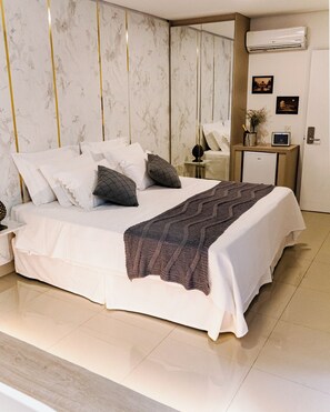 A modern hotel room with a large bed, a bedside table with a lamp, a small fridge, and a wall-mounted air conditioner. - Liguori Hotel (Trindade)