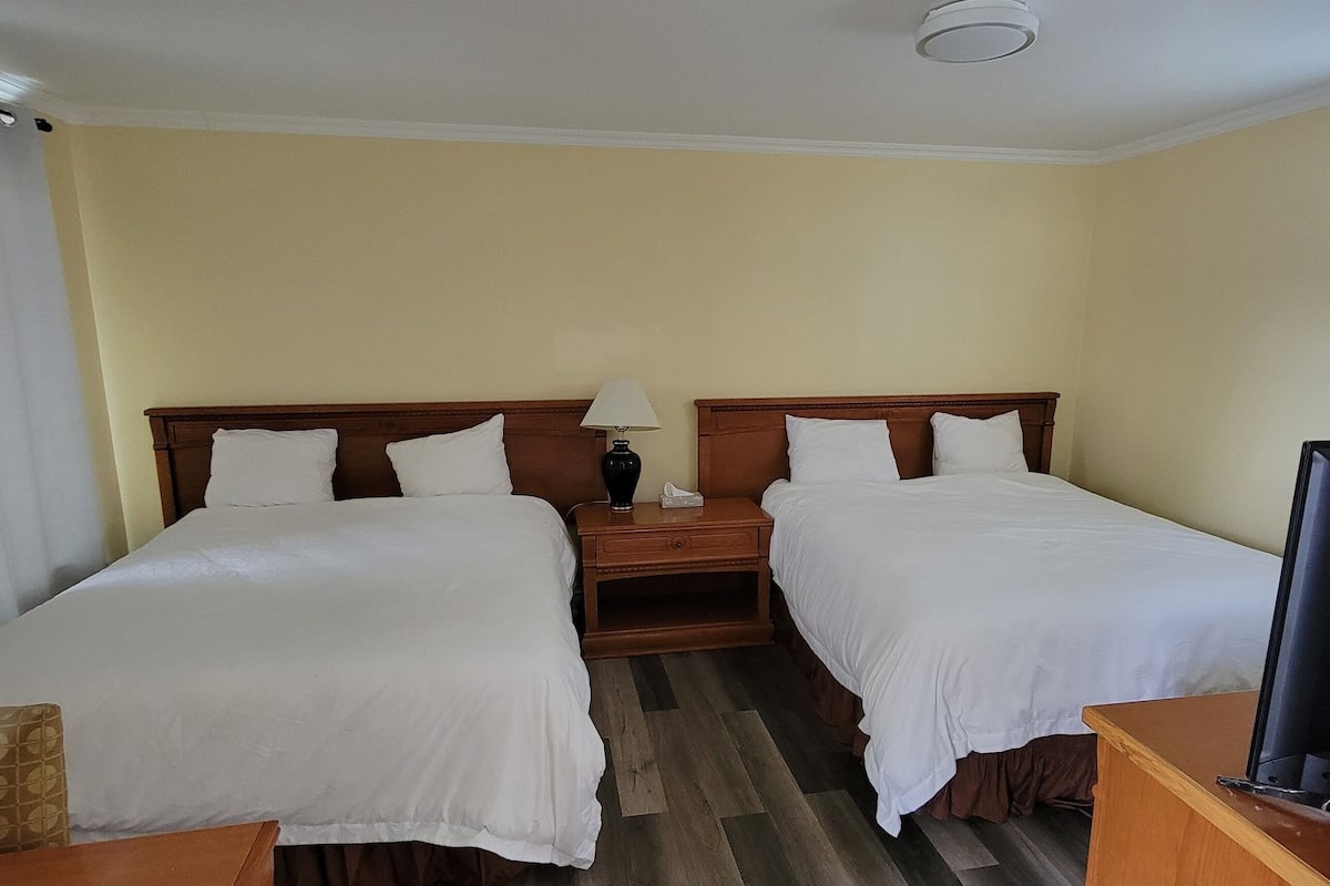 A hotel room with two beds, a nightstand, a lamp, and a television.