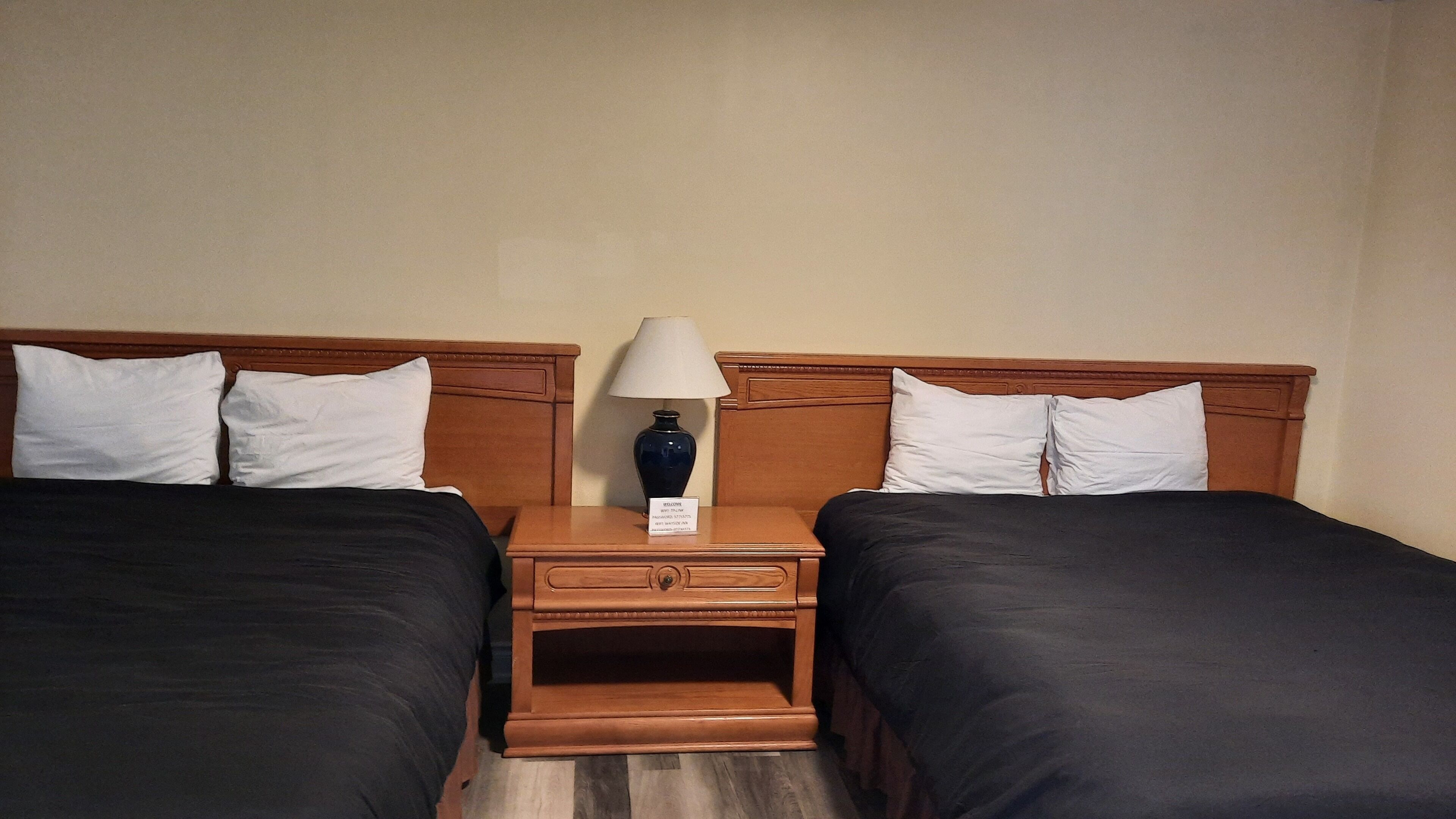 A hotel room with two beds, a nightstand with a lamp, and a welcome sign.