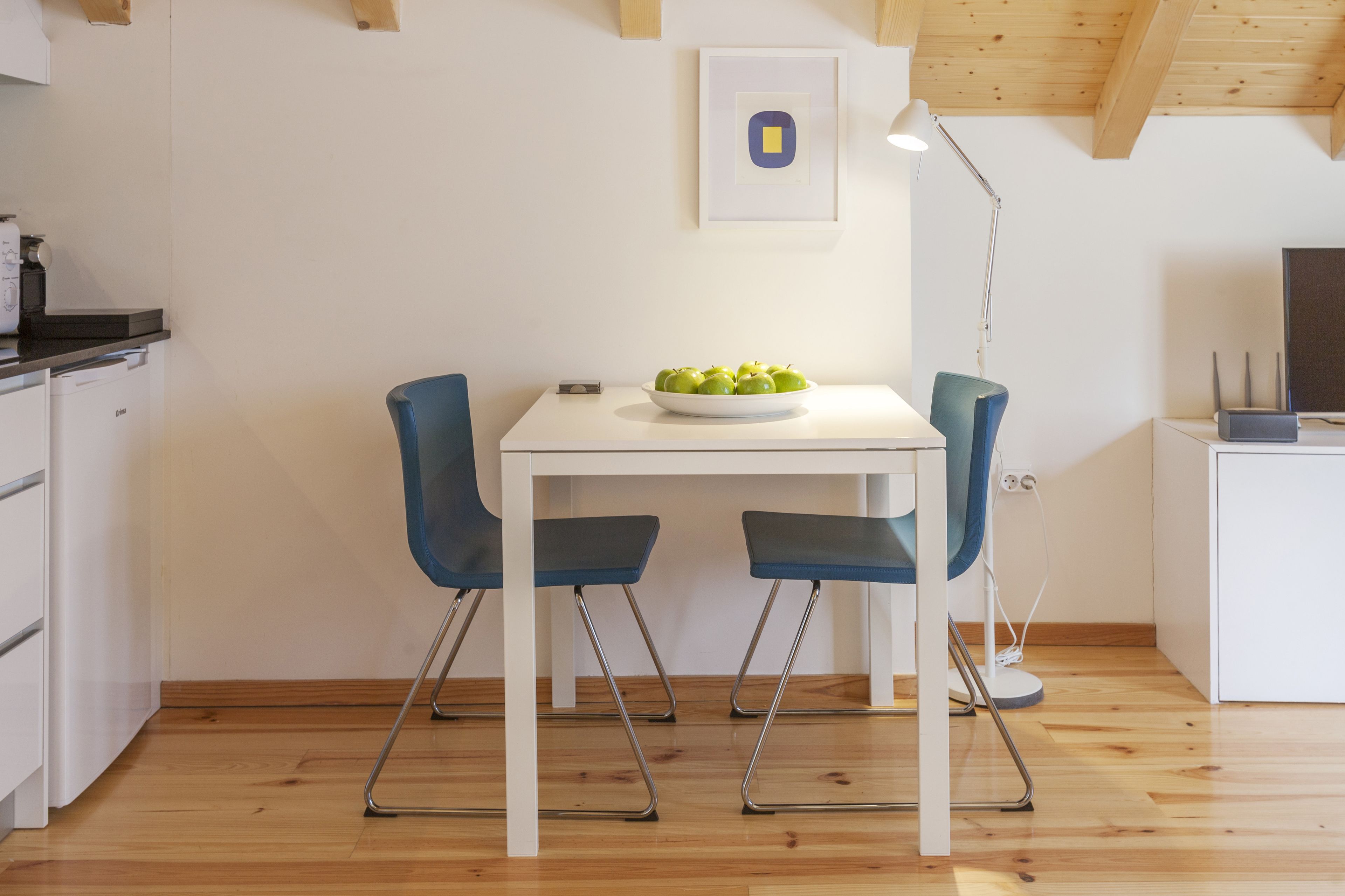 A modern kitchen with white cabinets, a white table, two blue chairs, and a bowl of green apples on the table.