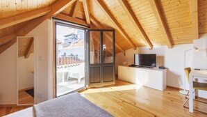 A modern, well-lit attic bedroom with wooden beams, a sliding glass door to a balcony, a bed, a TV, and a small white cabinet. - BO - Almada 340 (Porto)
