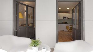 A modern, minimalist room with two white chairs, a small round table with a potted plant, and a door leading to a kitchen area with wooden flooring. - BO - Almada 340 (Porto)