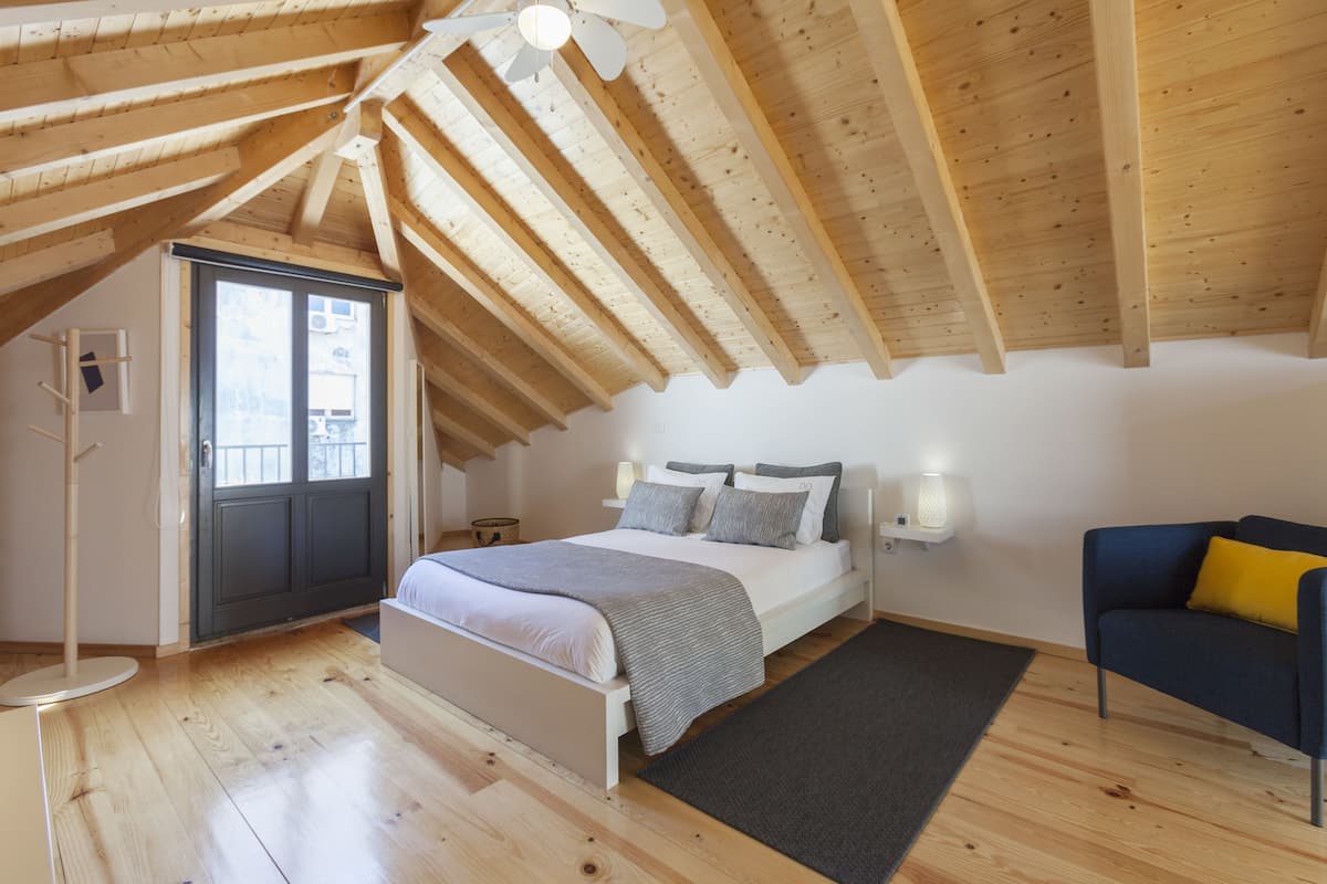 A bedroom with a sloped ceiling, a bed, bedside lamps, a chair, and a window.