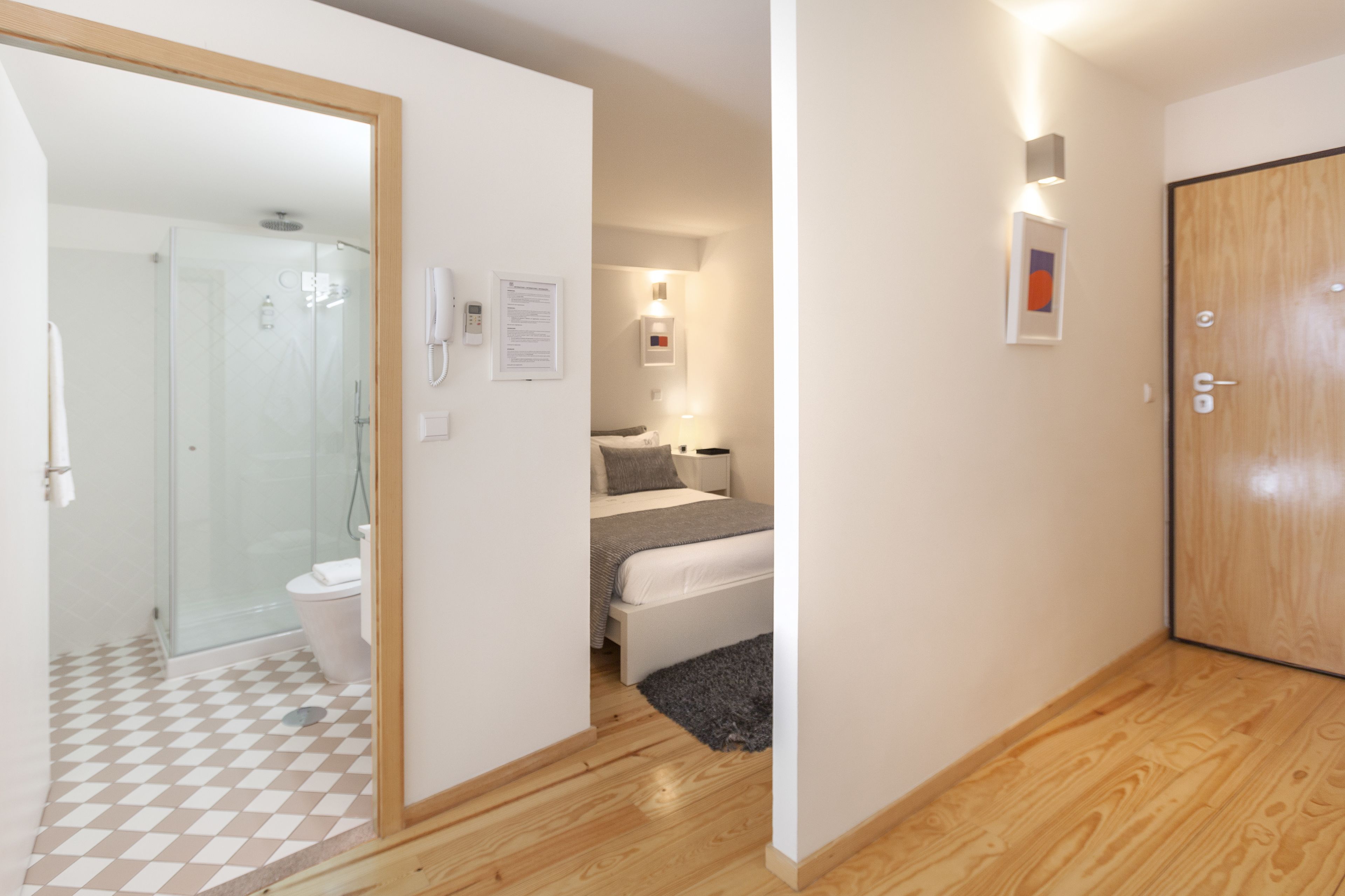 A modern hotel room with a glass shower enclosure, a bed with a gray blanket, and a wooden door.