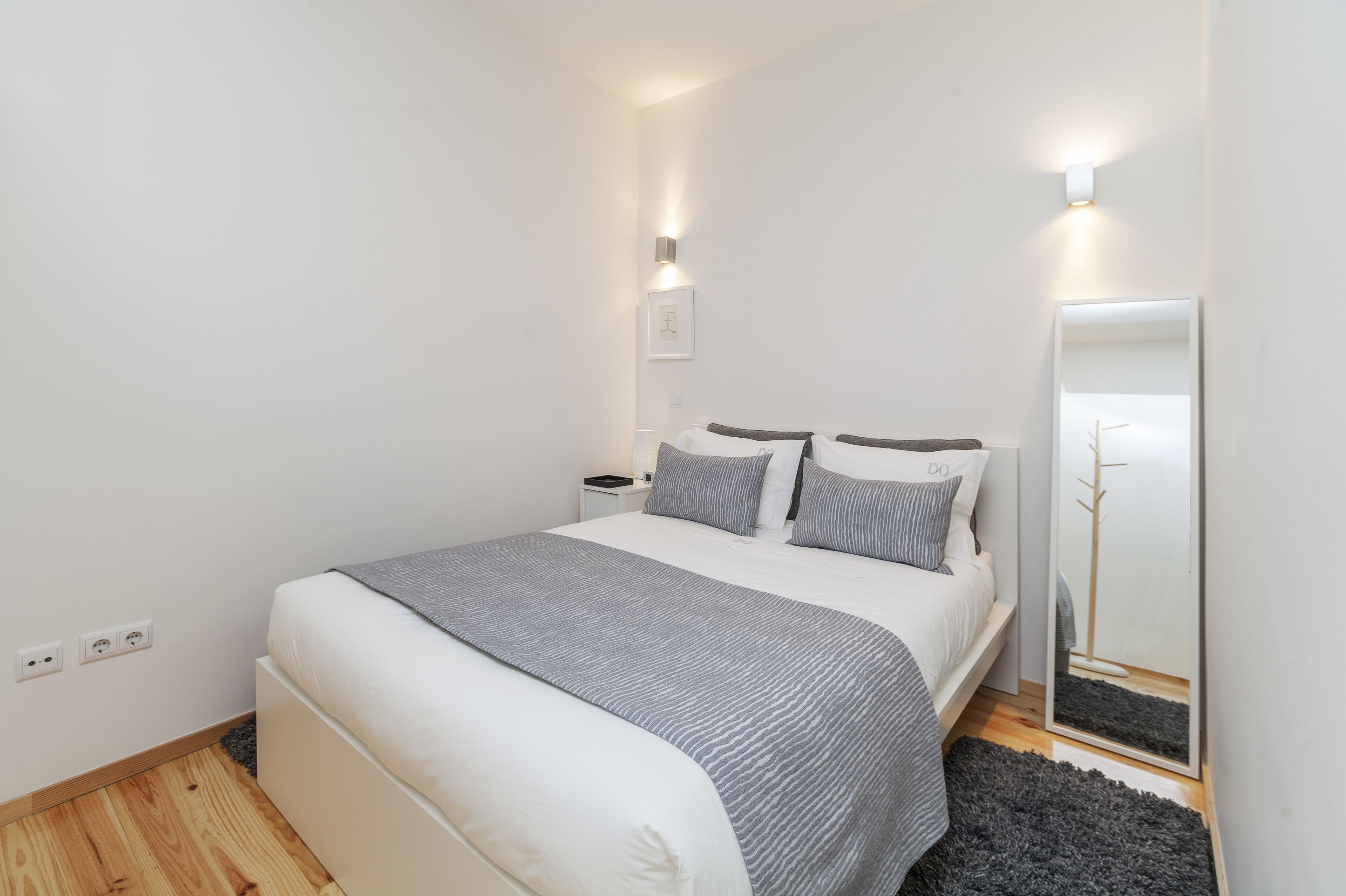 Un dormitorio ordenado con cama, mesitas de noche, un espejo y una lámpara fijada a la pared.