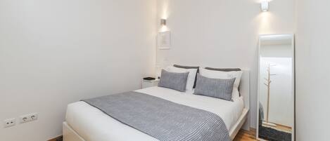 Un dormitorio ordenado con cama, mesitas de noche, un espejo y una lámpara fijada a la pared.