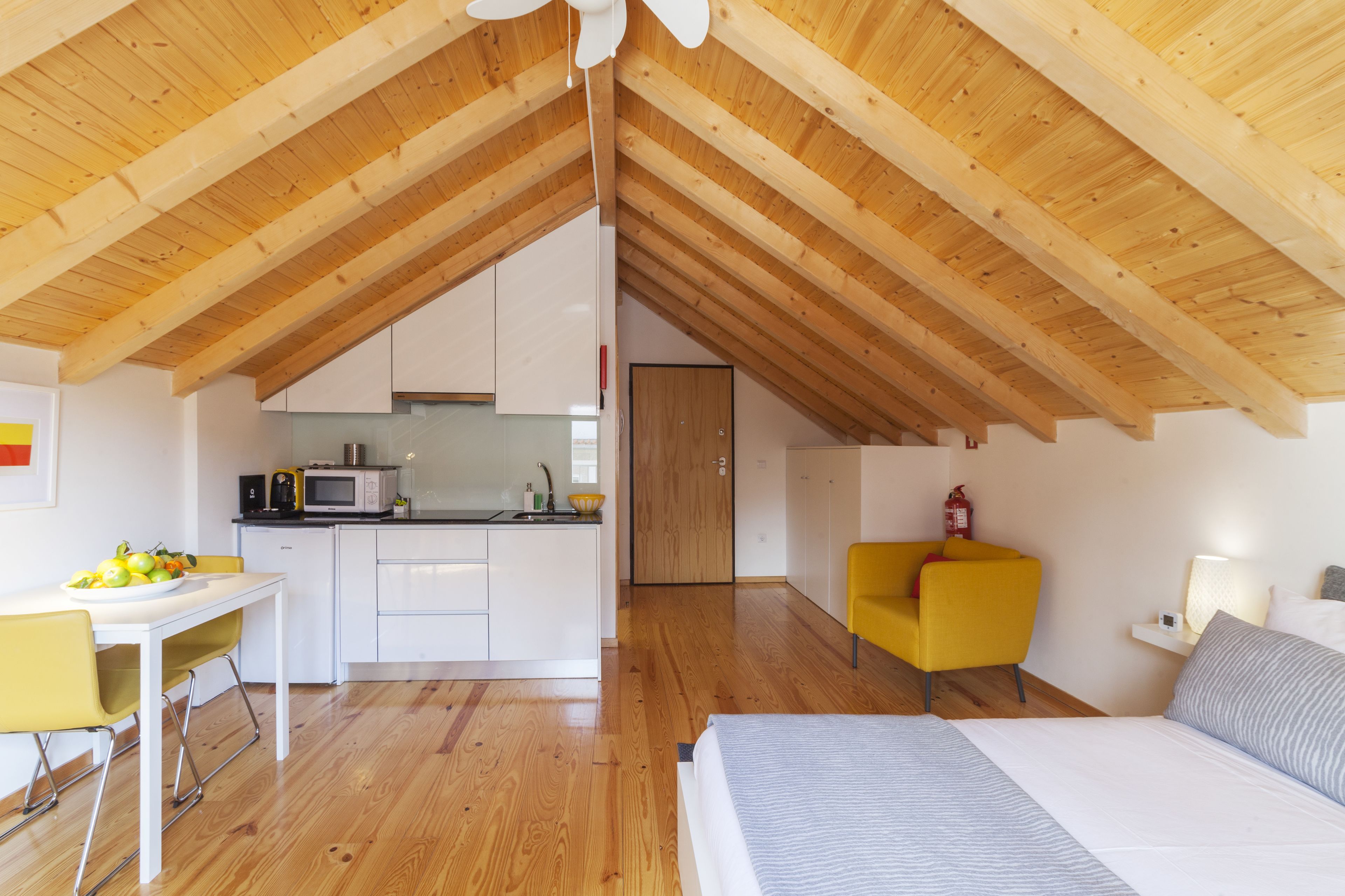Un dormitorio tipo loft moderno con techo inclinado, vigas de madera, una cama, un sillón amarillo y una kitchenette al fondo.