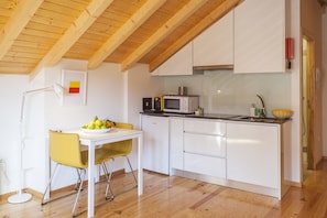 Una cocina moderna con armarios blancos, un microondas y un lavamanos. Hay una silla amarilla y una mesa blanca con fruta encima.
