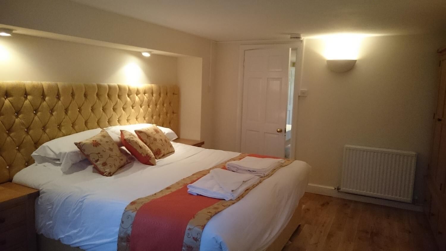 Un dormitorio con cabecera capitonada, cama con sábanas blancas y una manta roja, un radiador y una puerta.
