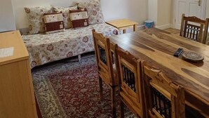 Een kleine, knusse kamer met een bed, een houten eettafel en stoelen. Aan de muur hangt een schilderij en er is een deur op de achtergrond te zien.