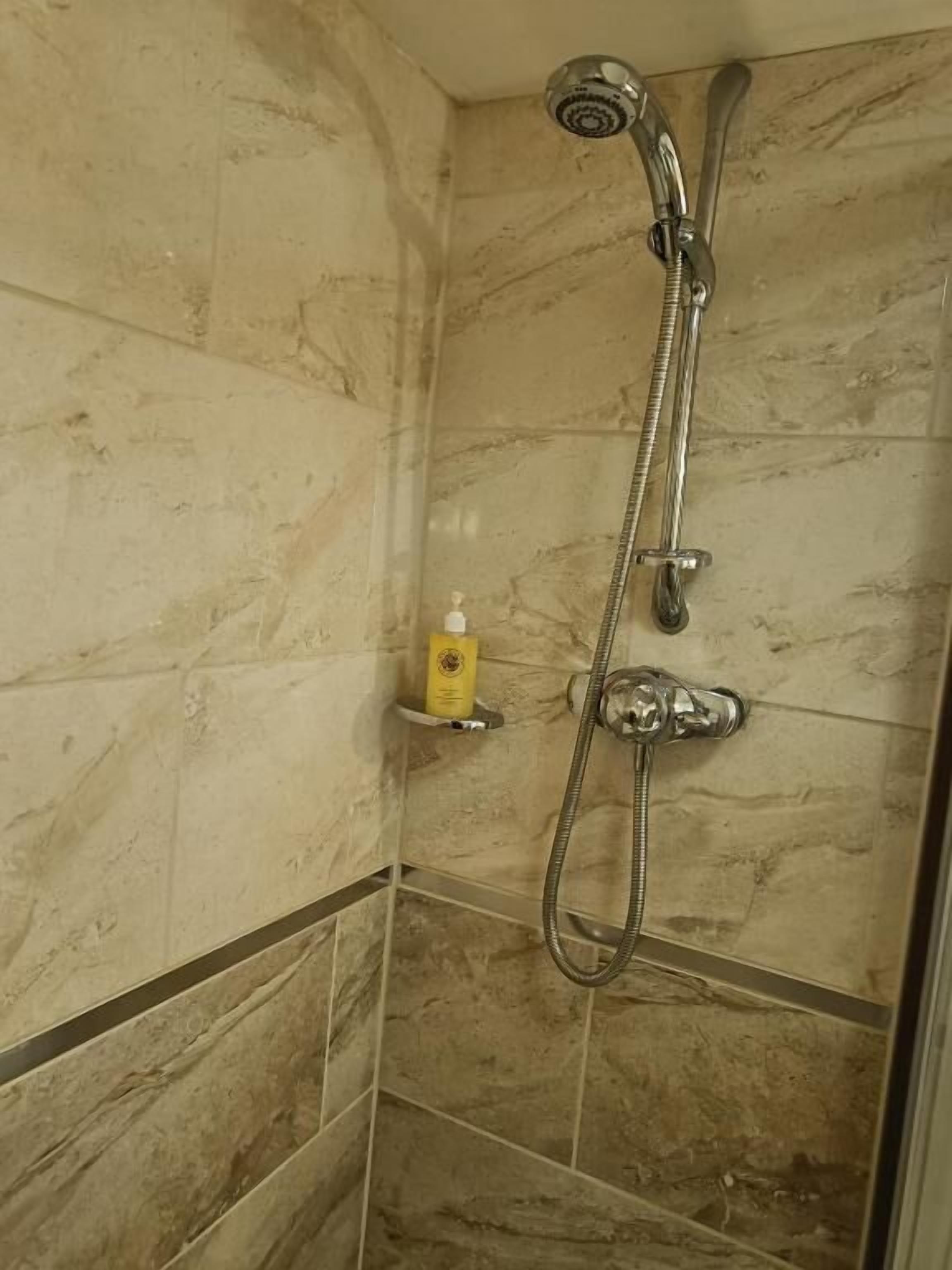 A shower with a handheld showerhead and a fixed showerhead, a yellow soap dispenser on a shelf, and tiled walls.
