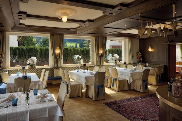 A dining area with tables set for a meal, chairs with blue sashes, and a view of greenery outside. - Hotel Savoy (Badia)