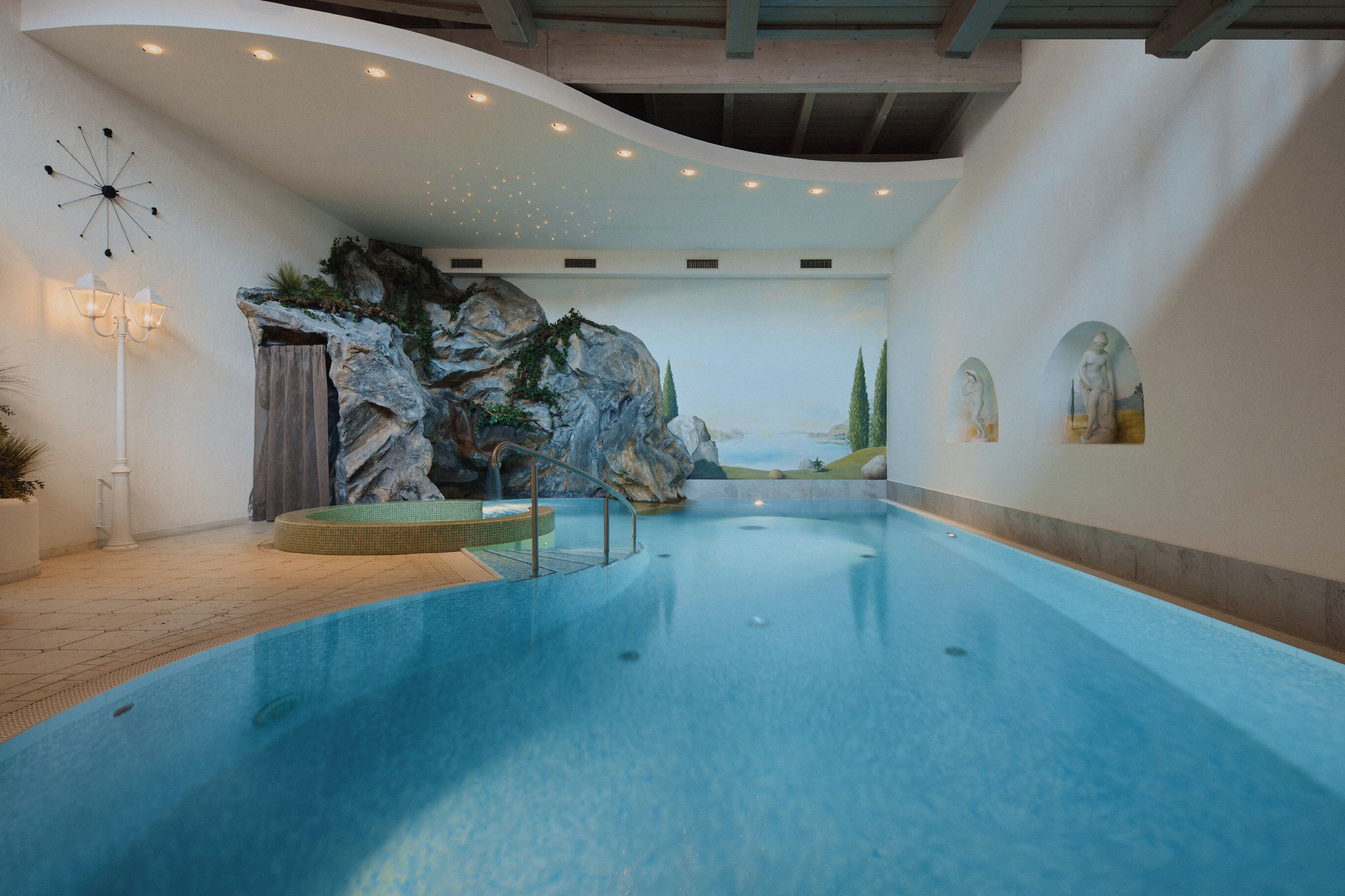 Una gran piscina interior con un mural de pared de rocas, un reloj y una estatua.