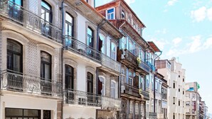 Una hilera de edificios de varios pisos con balcones, una calle con coches aparcados y cielo despejado.