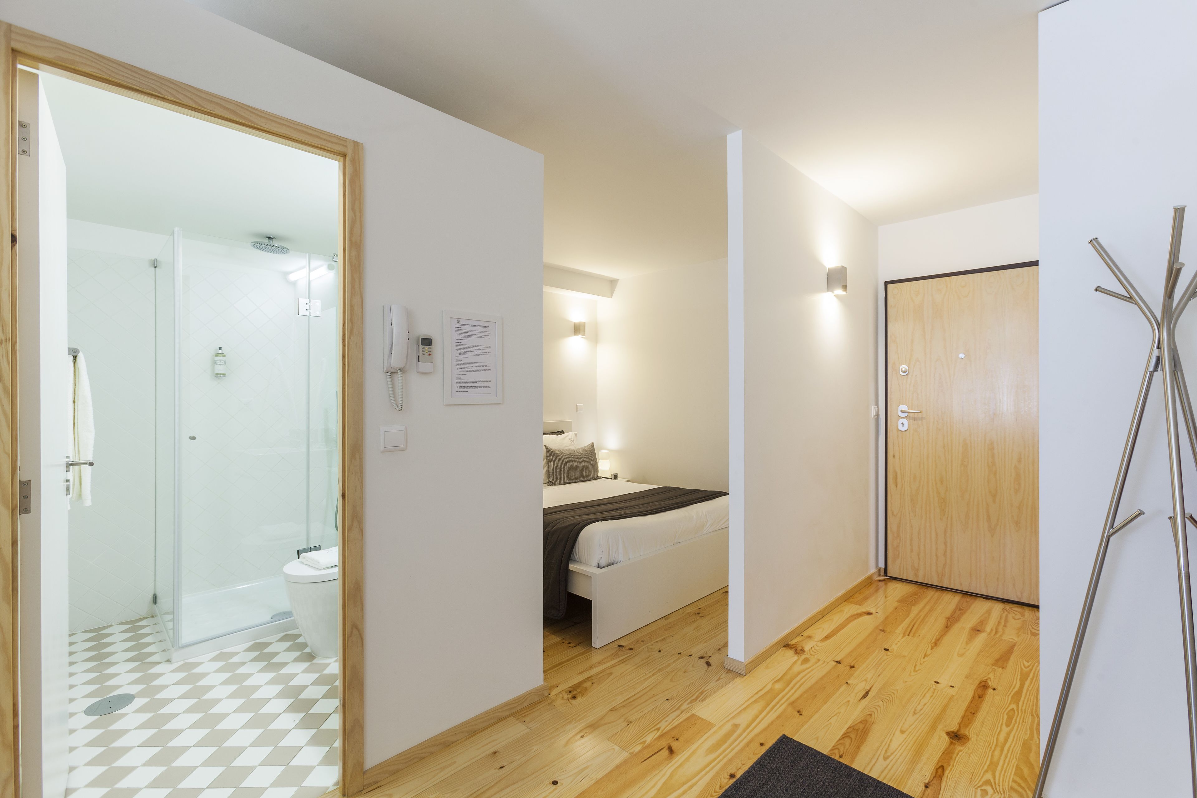 Habitación de hotel moderna con ducha de vidrio, piso de madera, cama con cabecera blanca y puerta de madera.