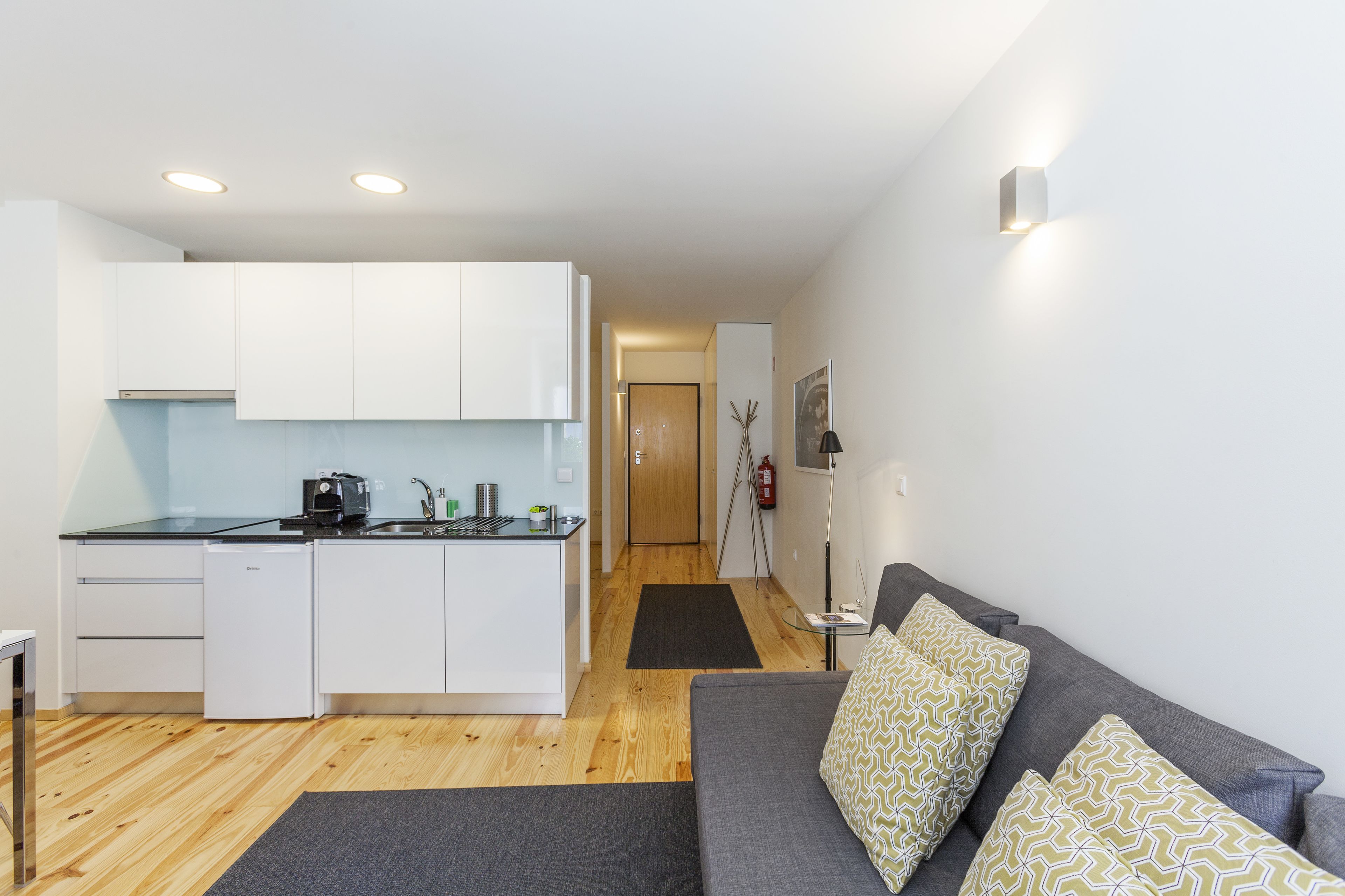 Un espacio habitable compacto con kitchenette, un sofá gris con cojines estampados y un pasillo que conduce a una puerta.