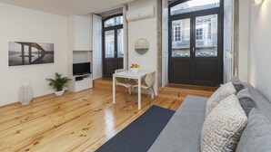 Una sala de estar moderna con piso de madera, un sofá blanco, una mesita con sillas, un televisor y un amplio ventanal con vista a edificios.