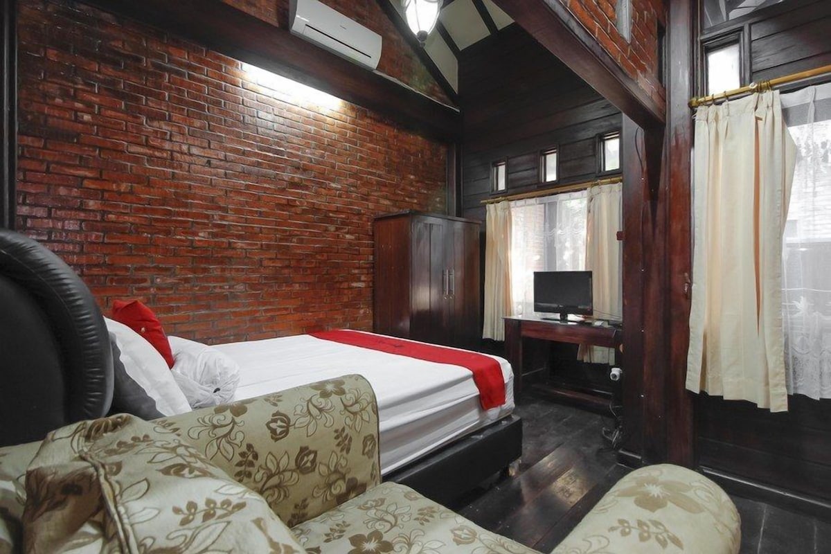 A room with a brick wall, a bed with a red and white bedspread, a wooden wardrobe, a television on a wooden stand, and a window with curtains.