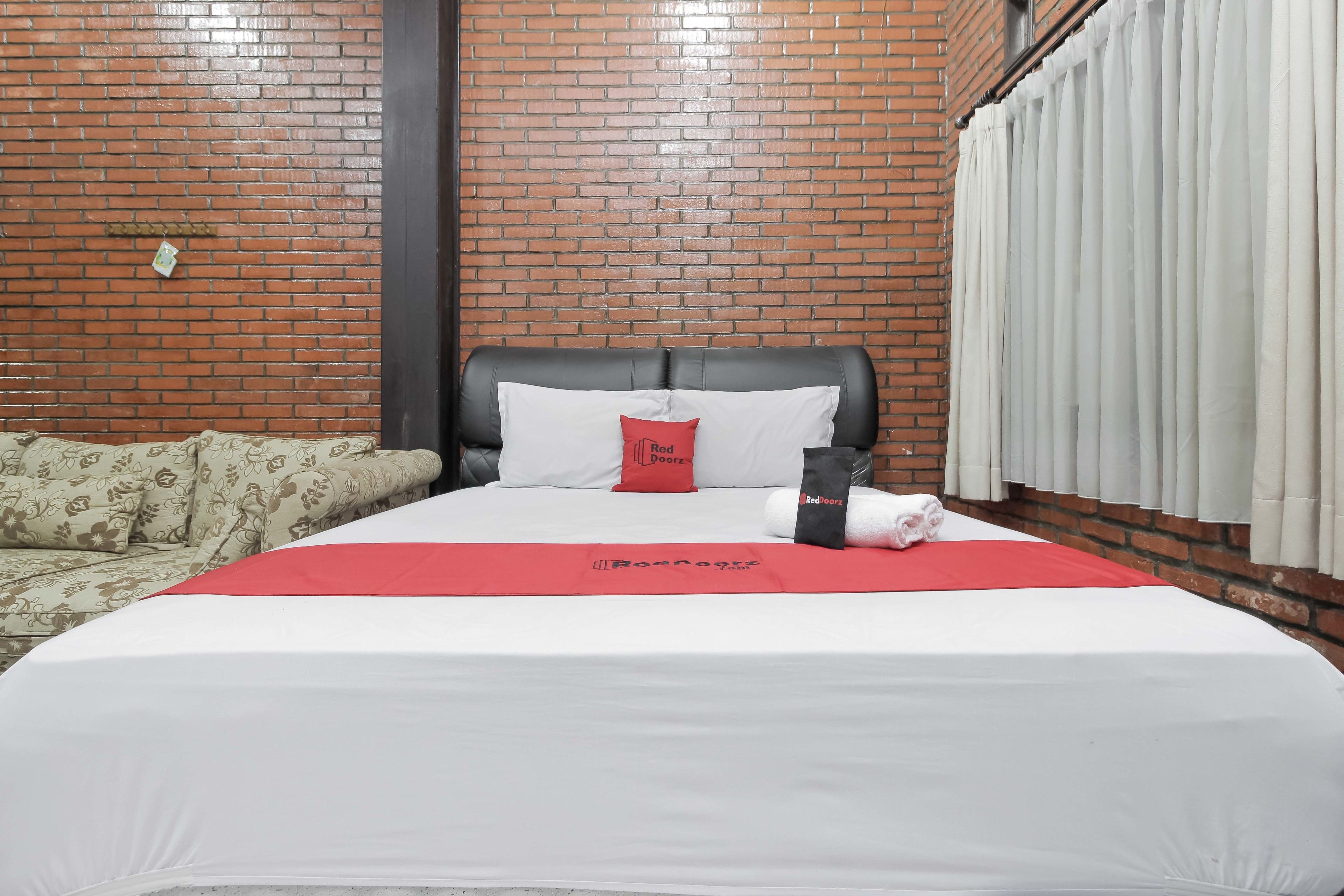 Hotelkamer met een bakstenen muur, een bed met wit beddengoed en een rood kussen, een zwart hoofdeinde en een witte gordijn.