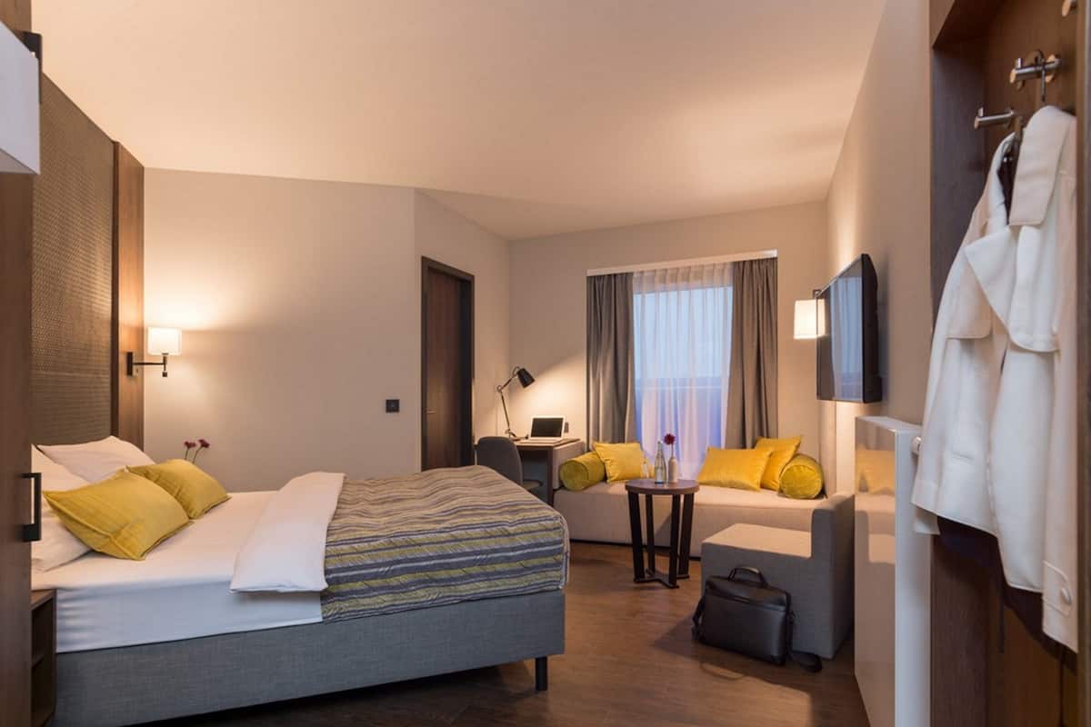 Una habitación de hotel con cama, sofá, escritorio con lámpara, televisor y perchero.