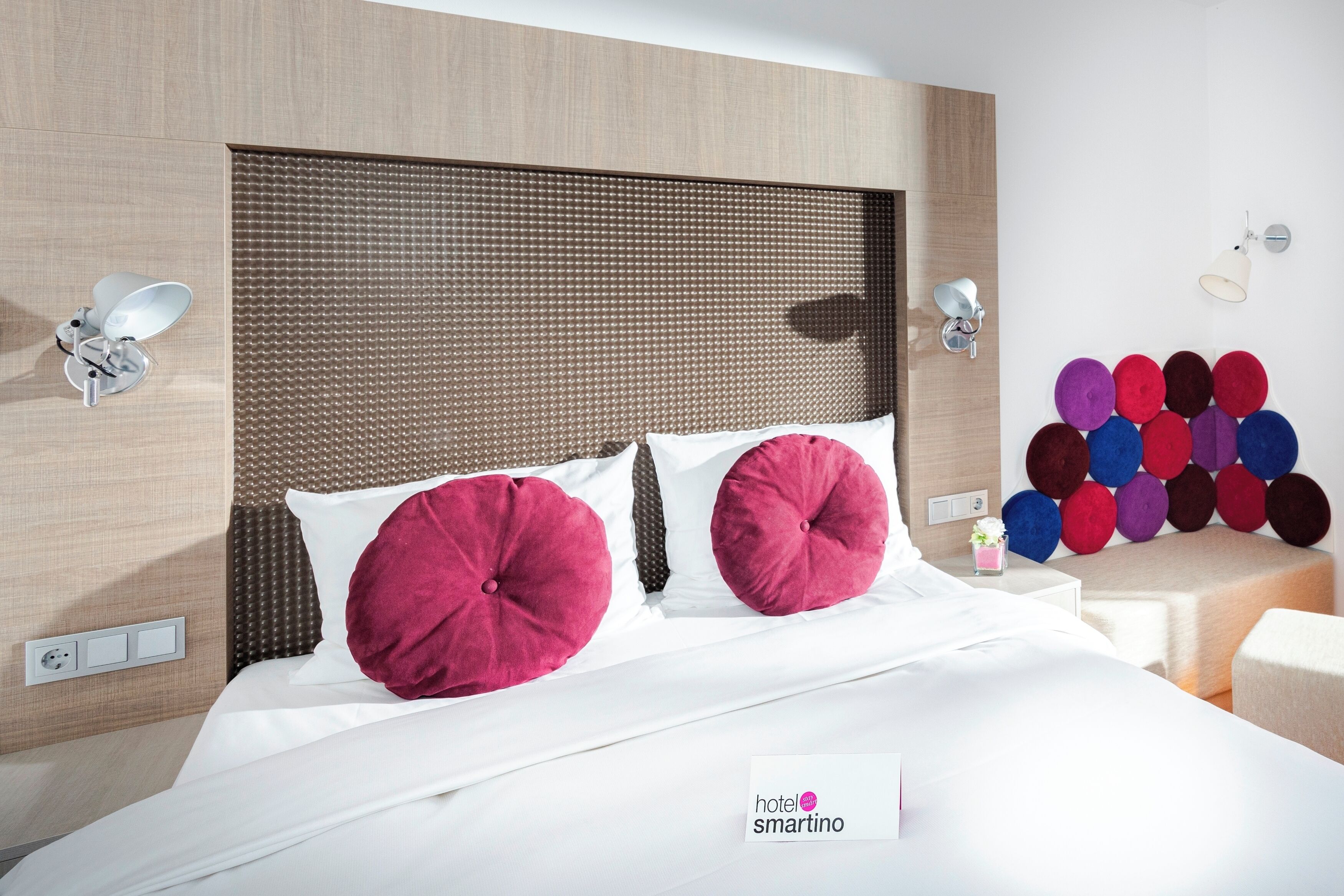 A hotel room with a bed, a headboard, wall-mounted lamps, and a wall decoration of colorful circles.
