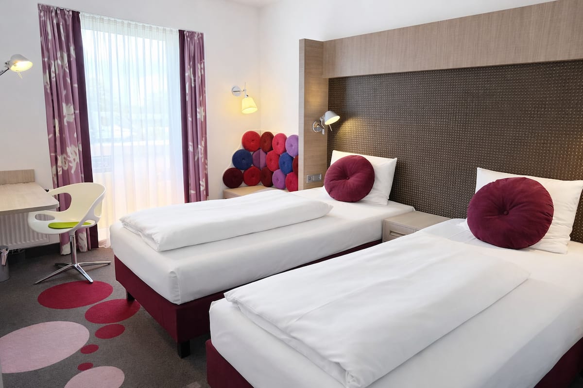 Habitación de hotel con dos camas, un escritorio, una silla, un radiador y una lámpara fijada a la pared.
