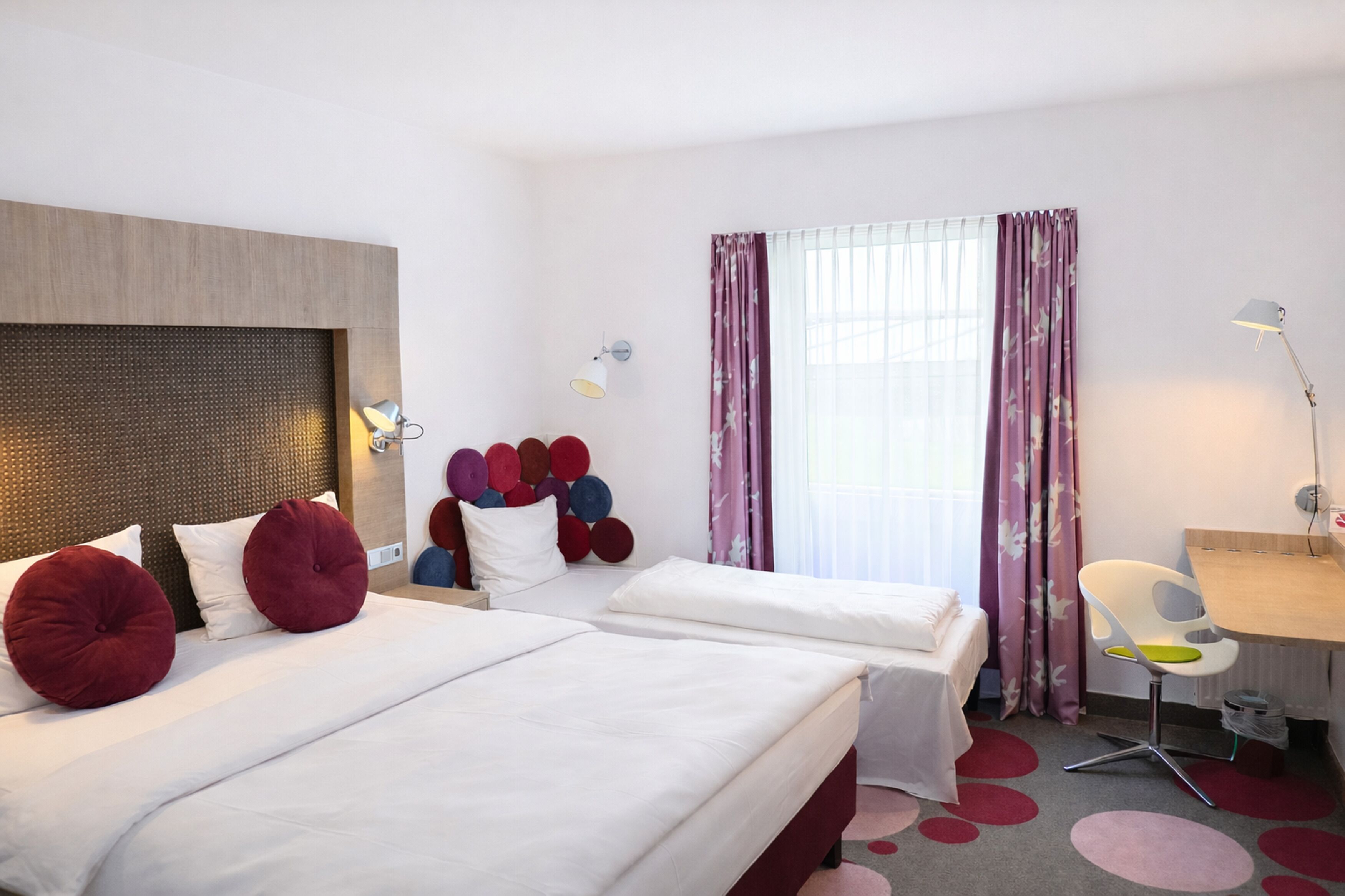 Une chambre d’hôtel avec deux lits, un bureau, une chaise, une lampe et une fenêtre avec des rideaux.