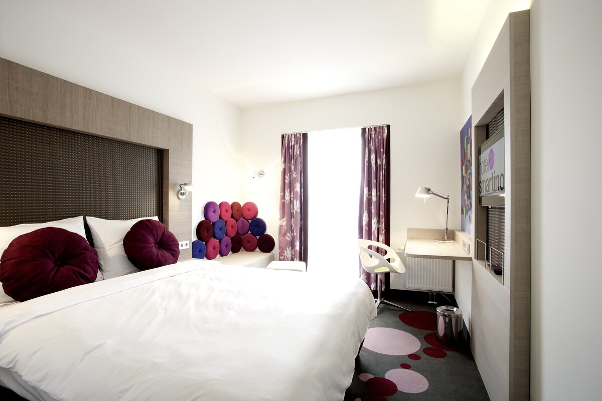 Una habitación de hotel con cama, escritorio, silla, radiador y cortinas.