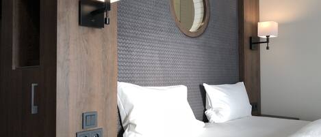 A hotel room with a bed, two bedside lamps, a round mirror, a nightstand with a remote control, and a wall-mounted switchboard.