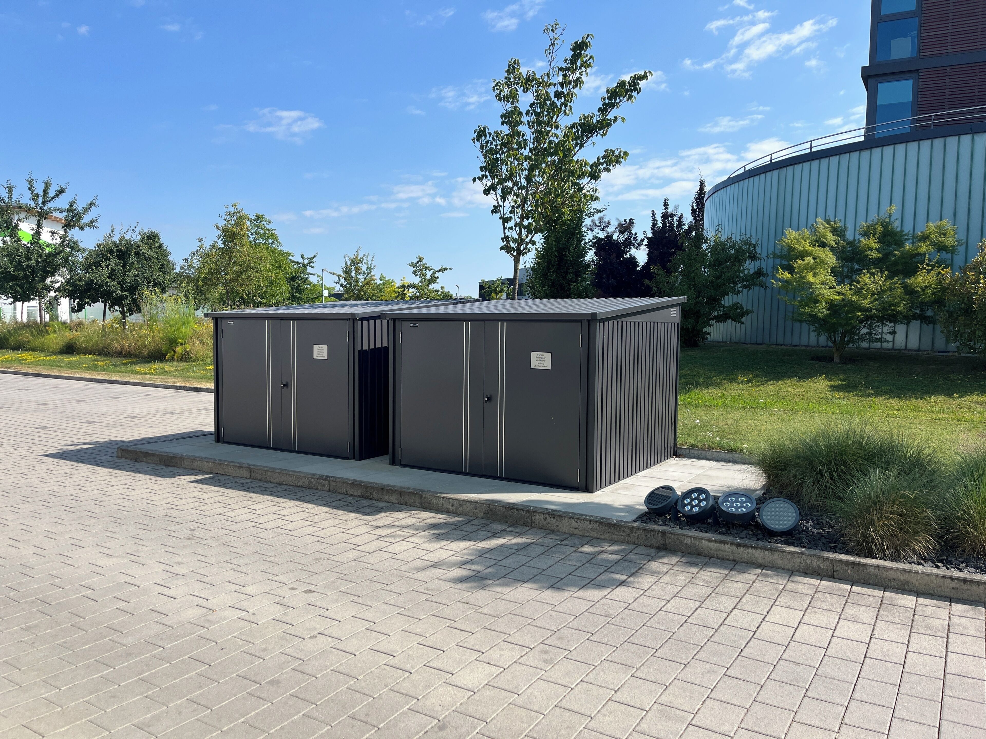 Un contenedor de almacenamiento portátil con dos puertas, ubicado en una zona pavimentada rodeada de césped y árboles.