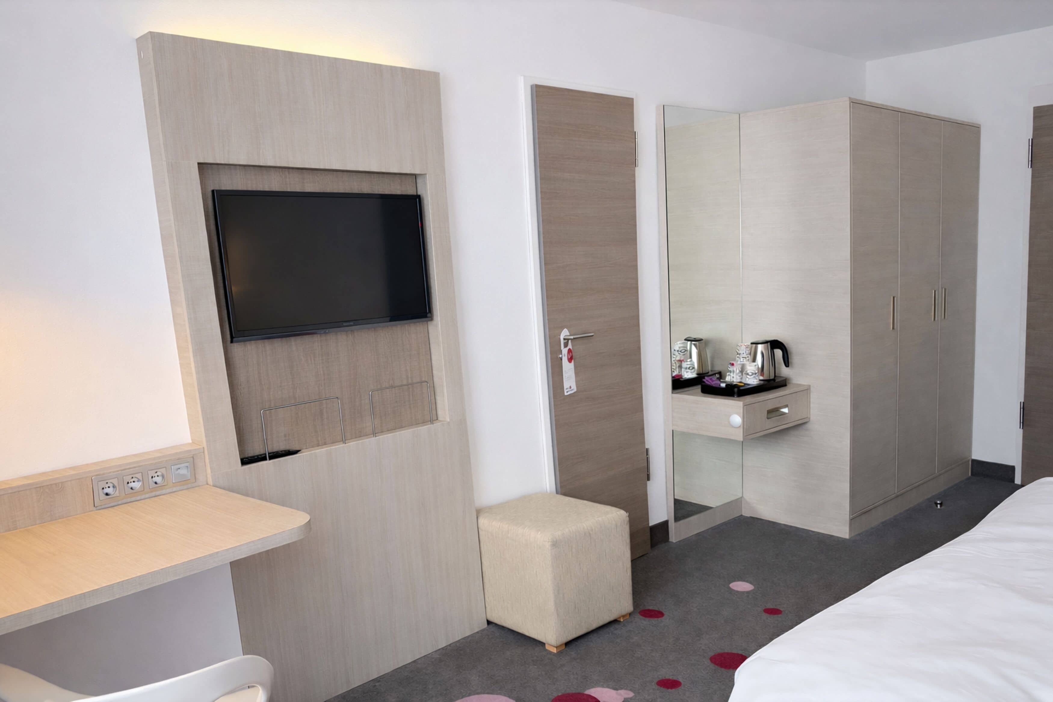 A hotel room with a flat-screen TV mounted on a wall, a small bedside table with a kettle, a wardrobe, and a bed with a white duvet.