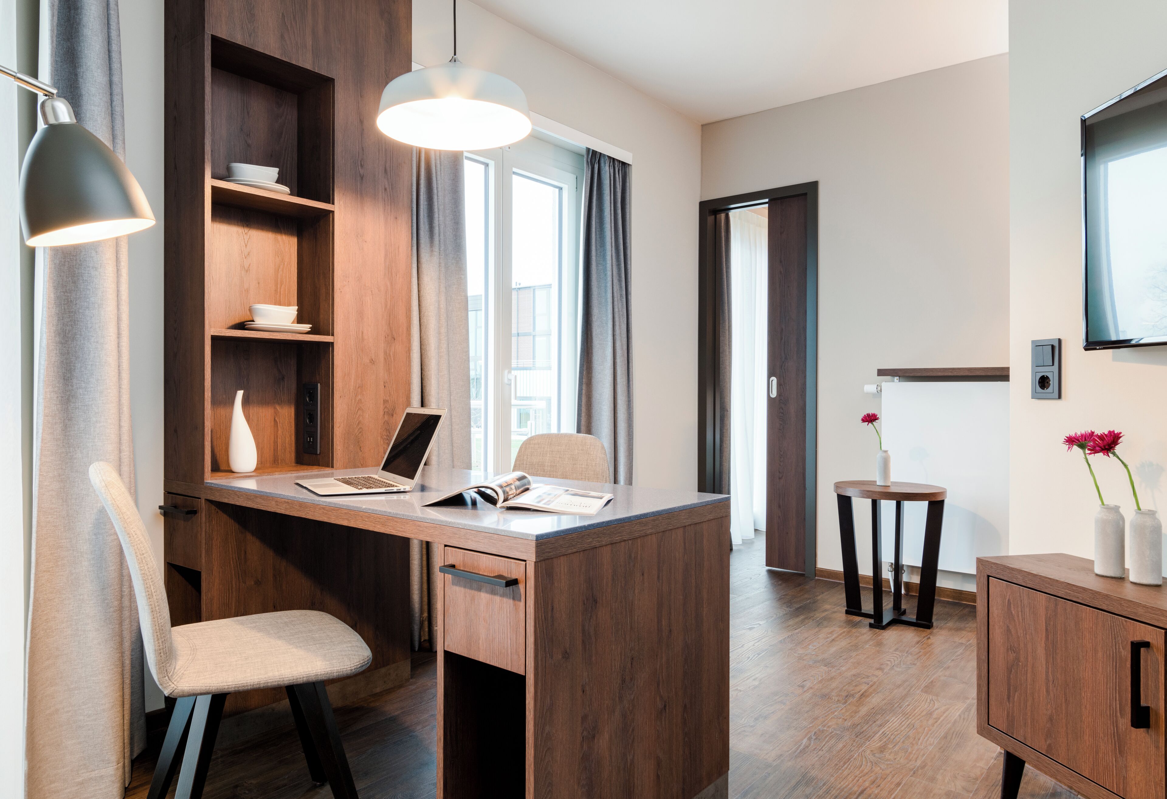 Una habitación moderna con un escritorio de madera, una silla, una computadora portátil, una mesita con un florero y un televisor montado en la pared.