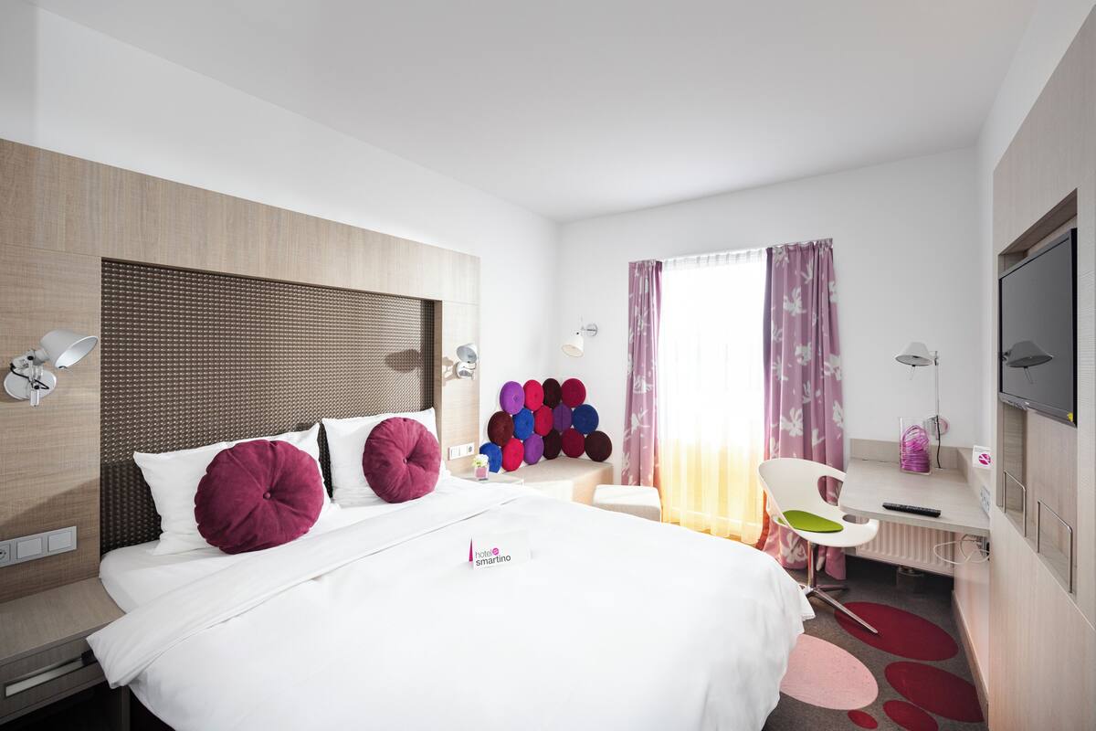 Una habitación de hotel con cama, escritorio, silla, televisor y lámpara de pared.