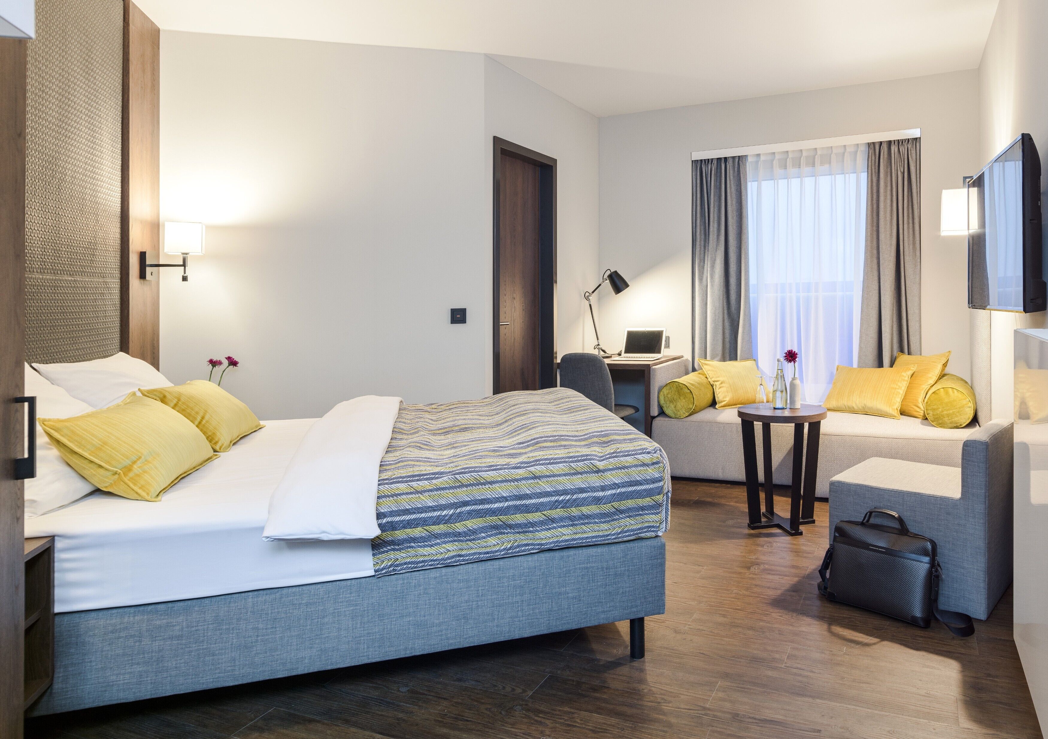 A hotel room with a bed, a sofa, a desk, a chair, a TV, and a lamp.