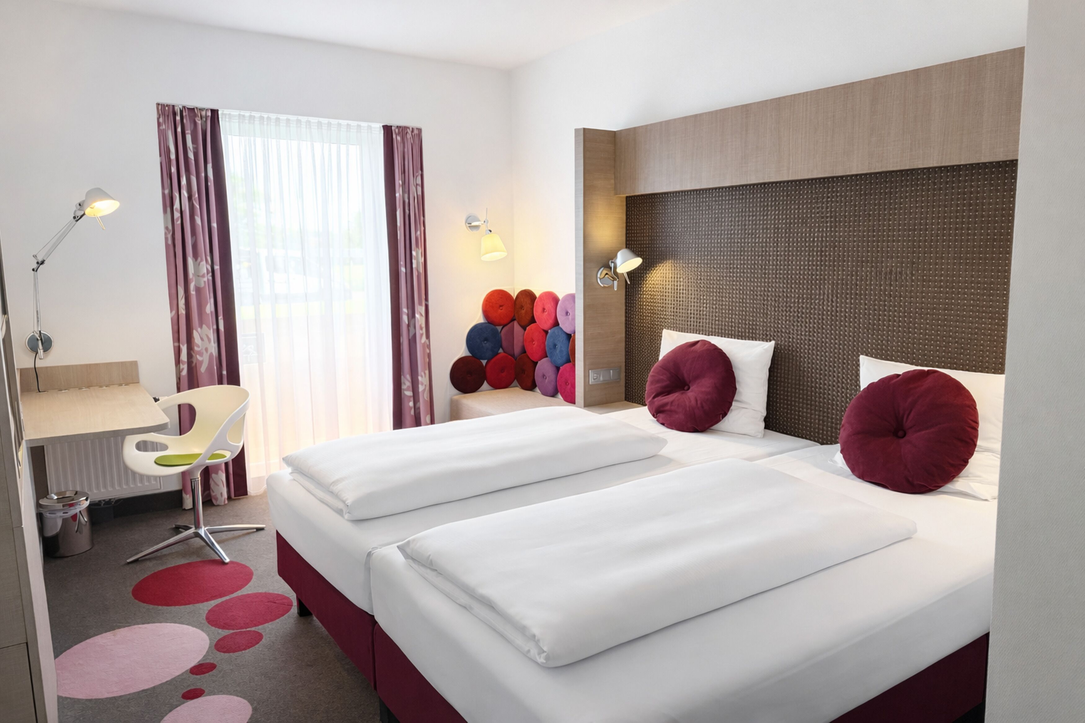 A hotel room with two beds, a desk, a chair, a radiator, and a wall with colorful circular decorations.