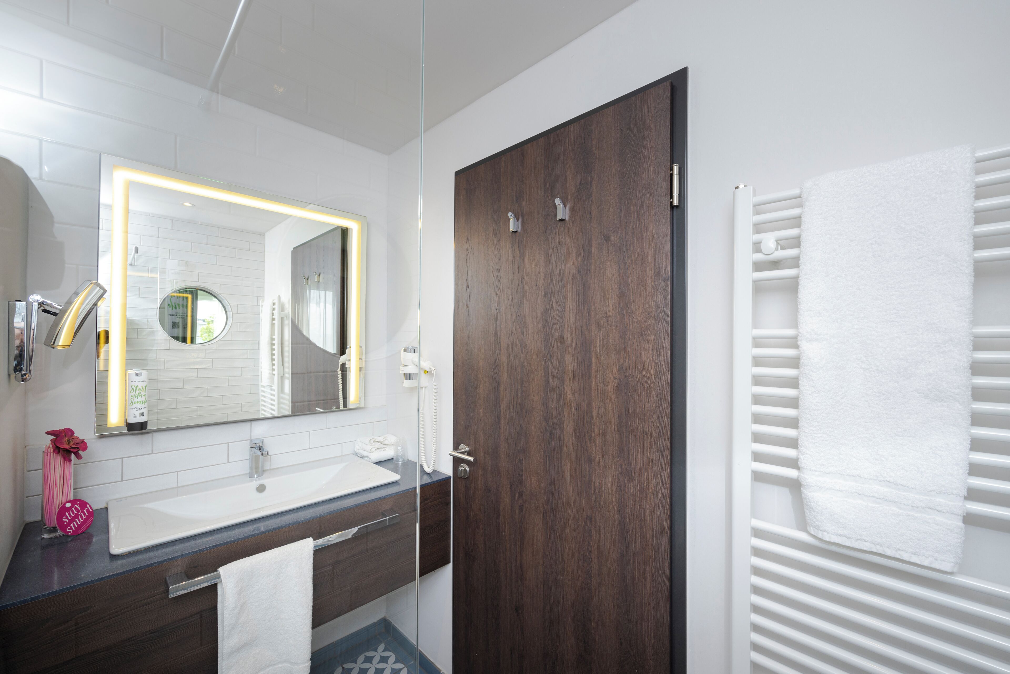 Een moderne badkamer met een grote spiegel, een witte wastafel, een houten deur en een handdoekrek.