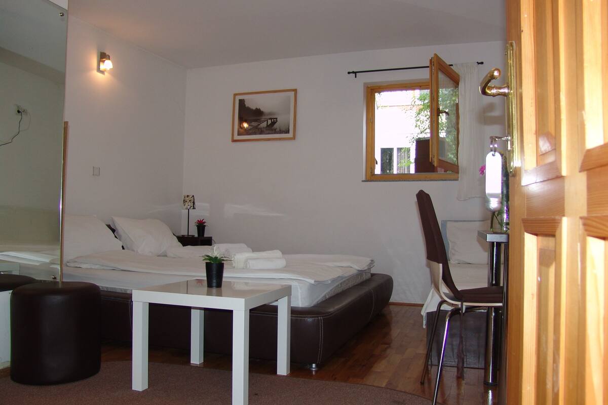 A bedroom with a bed, a small table, a chair, a bedside table with a lamp, a picture on the wall, and a window with a view of greenery.