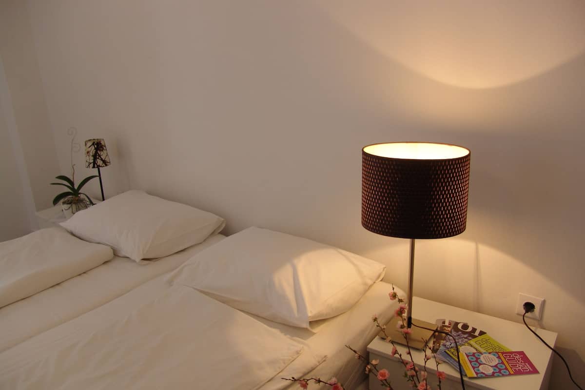 A neatly made bed with white linens, a bedside lamp with a patterned shade, and a small table with a plant and a few books.