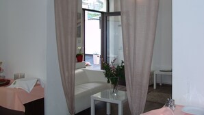 Ein Zimmer mit einer weißen Couch, einem kleinen Tisch mit Blumenvase und einem Fenster mit Vorhängen.