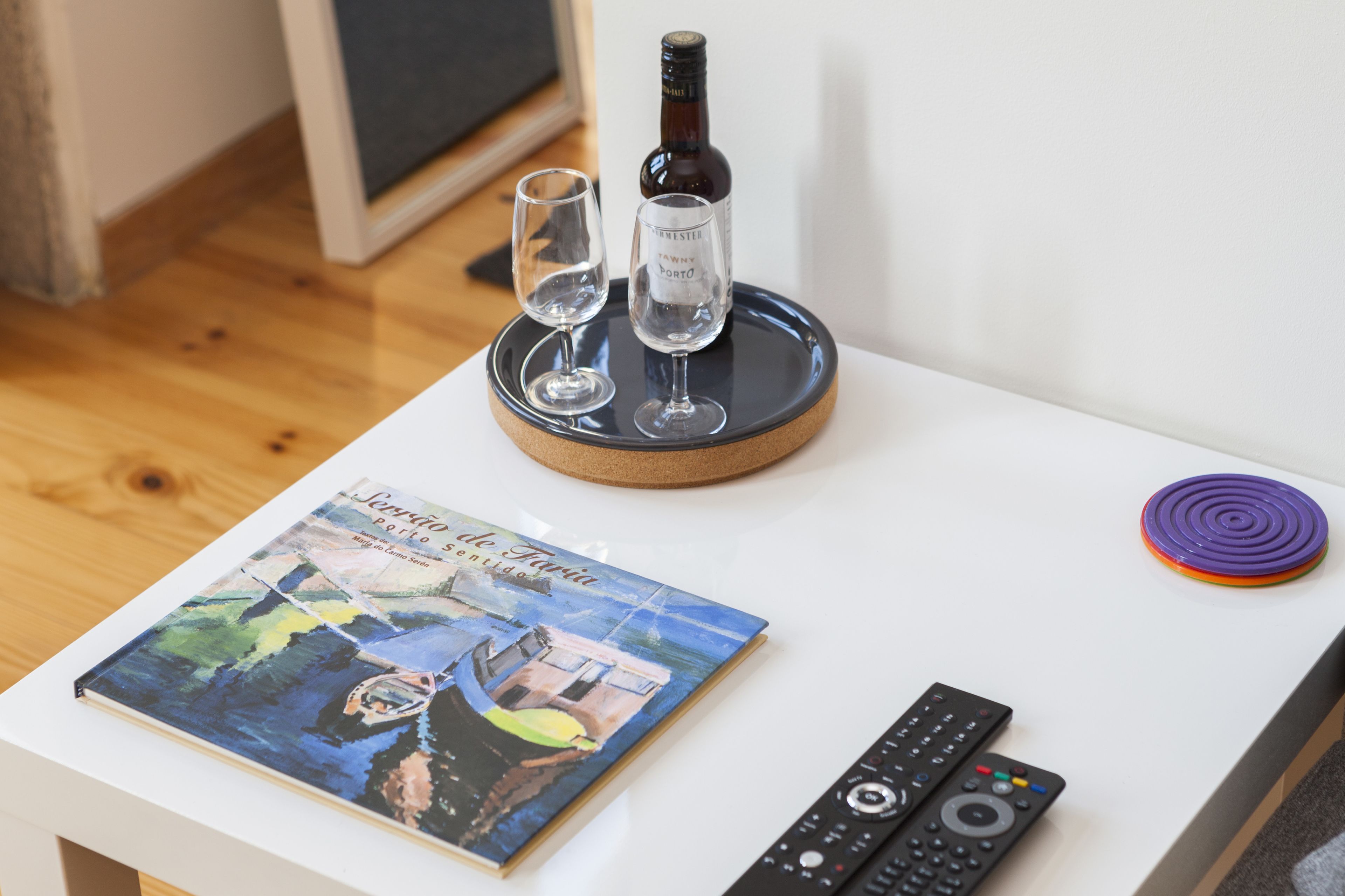 A table with a cork coaster holding a bottle and two glasses, a book, and two remote controls.