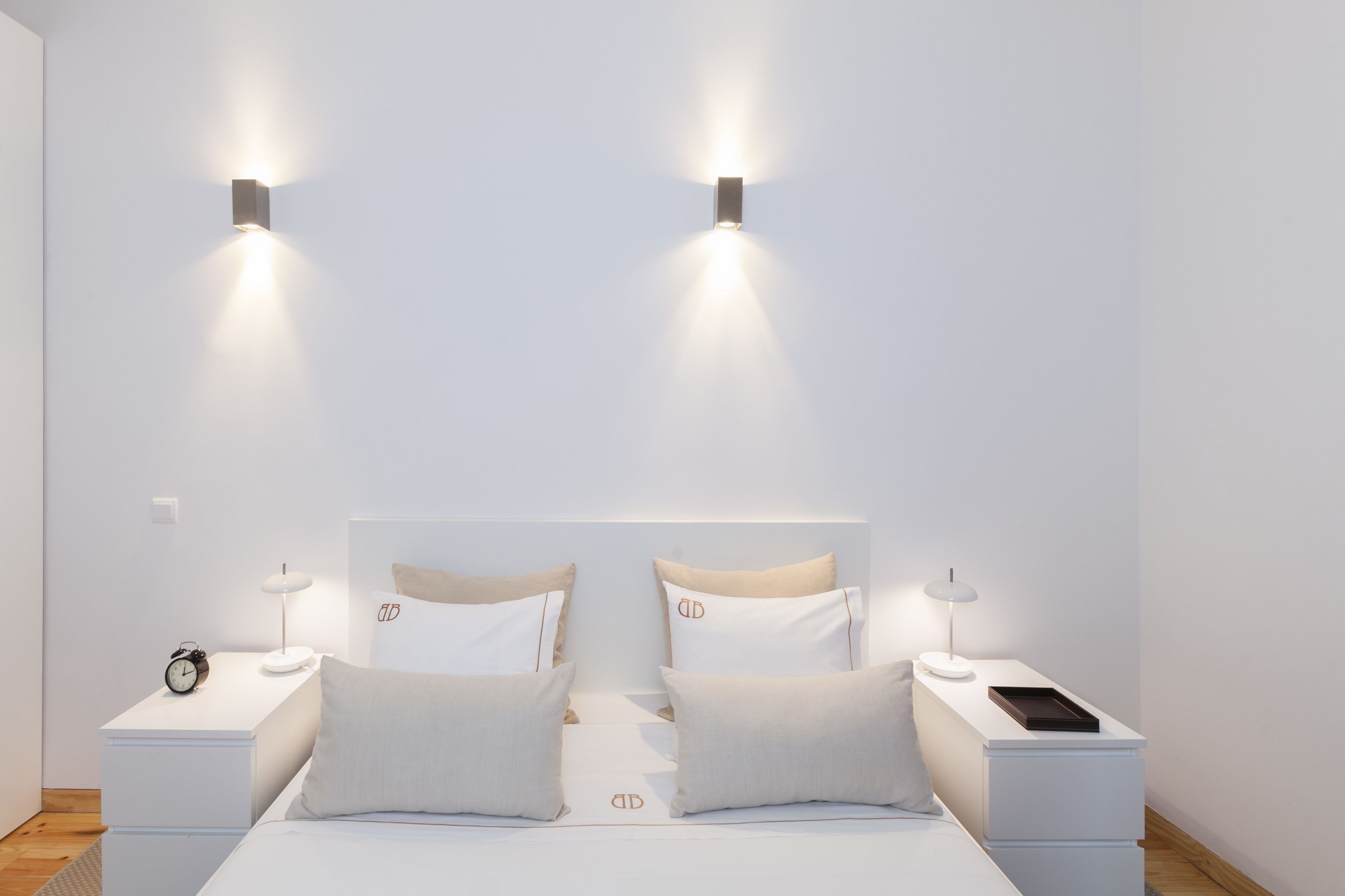 A modern bedroom with a large bed, two bedside tables, a nightstand, a wall-mounted light fixture, and a small clock.