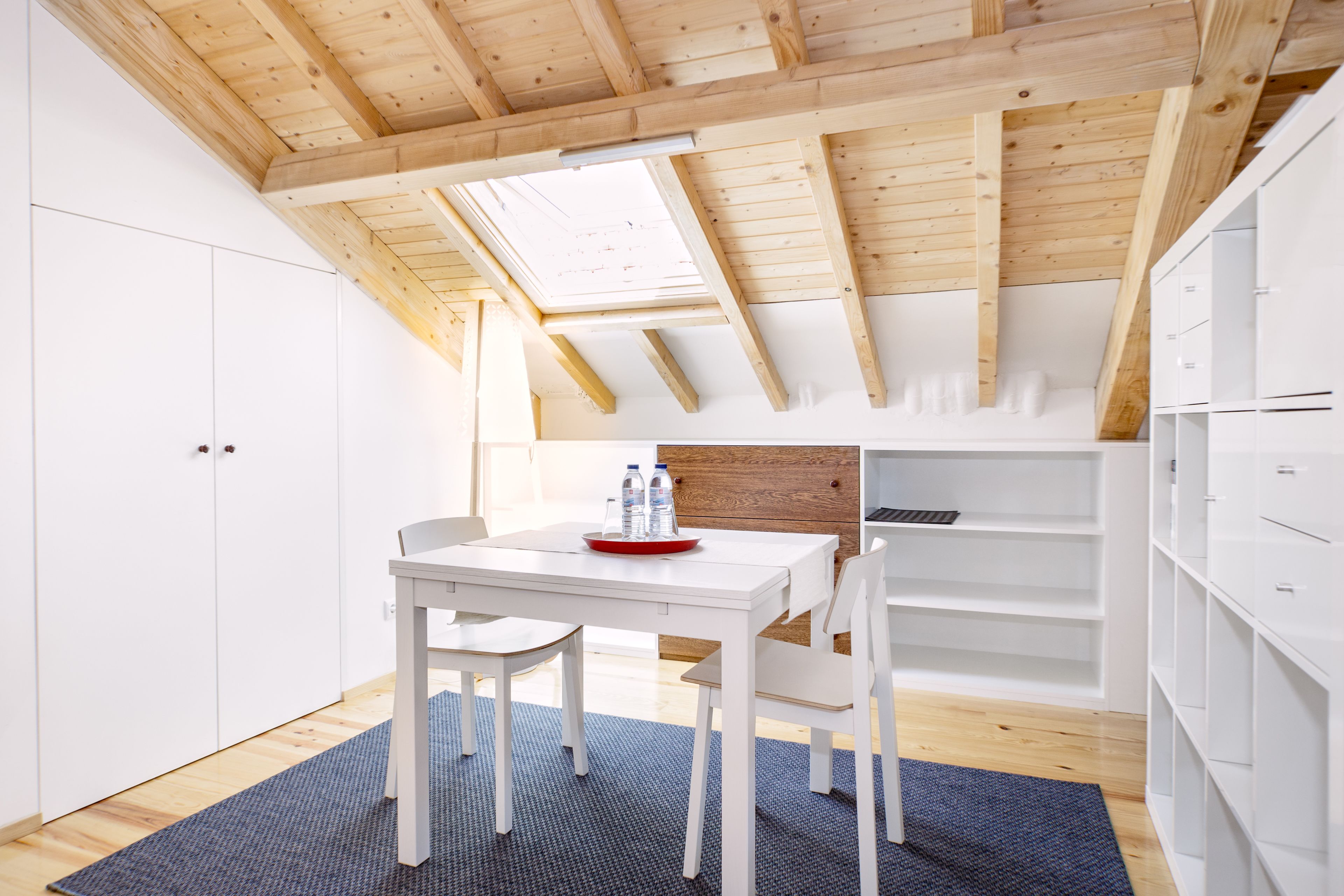 A modern, well-lit attic room with a sloped ceiling, white walls, and a skylight. It features a white dining table with chairs, a blue rug, and built-in white shelving.