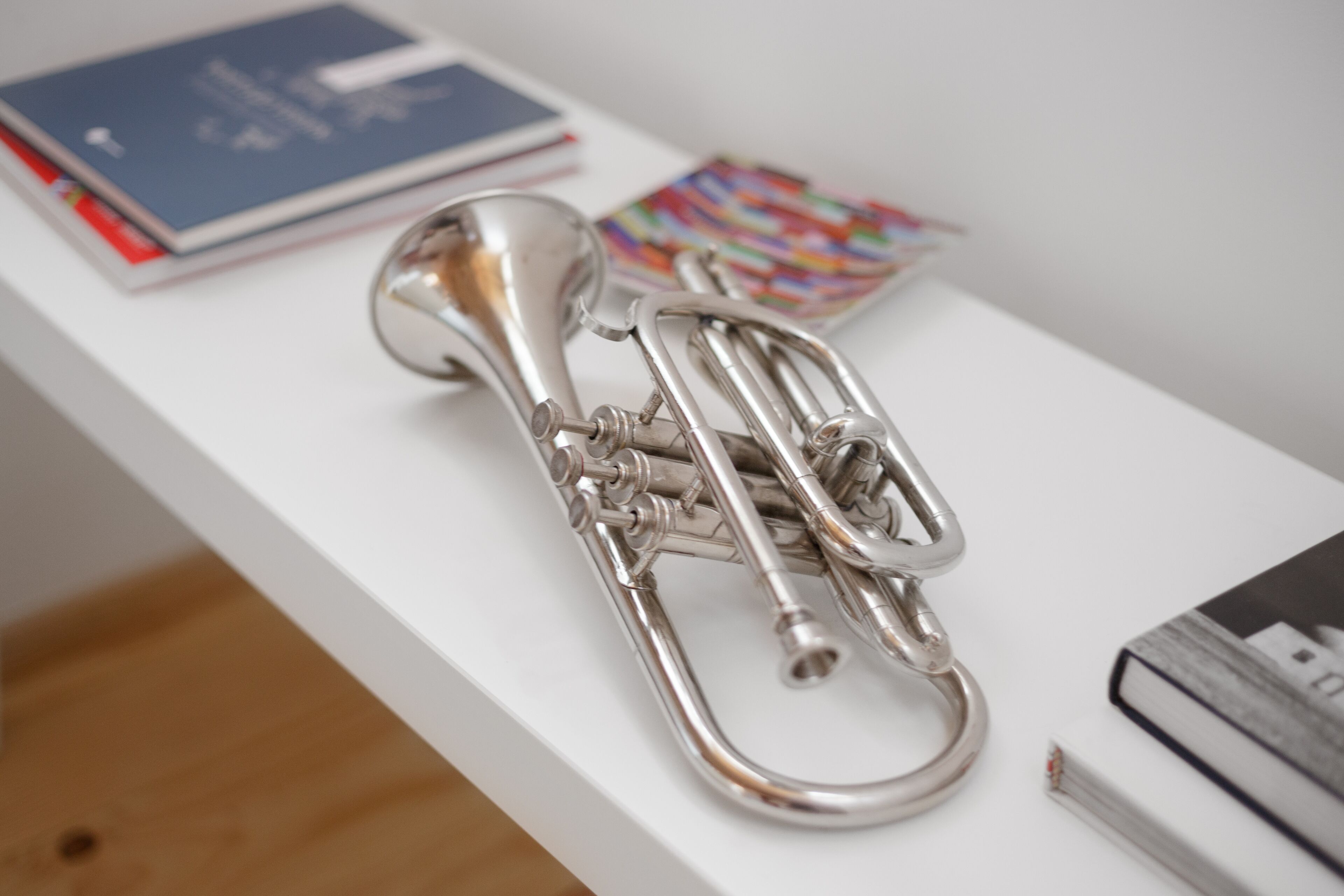 A silver trumpet on a white surface with books and a colorful item in the background.