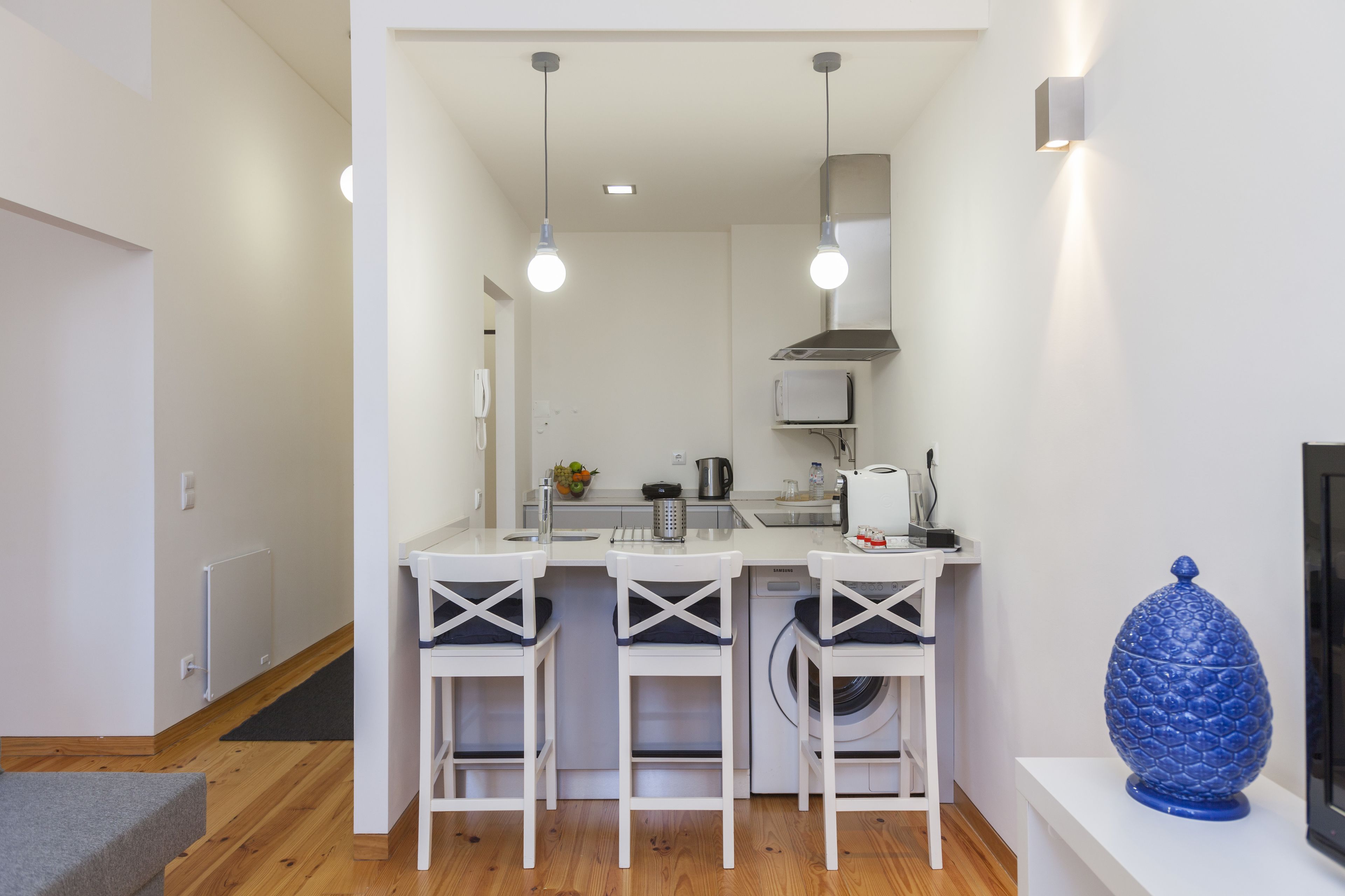 A modern kitchen with white bar stools, a built-in washing machine, and pendant lighting.