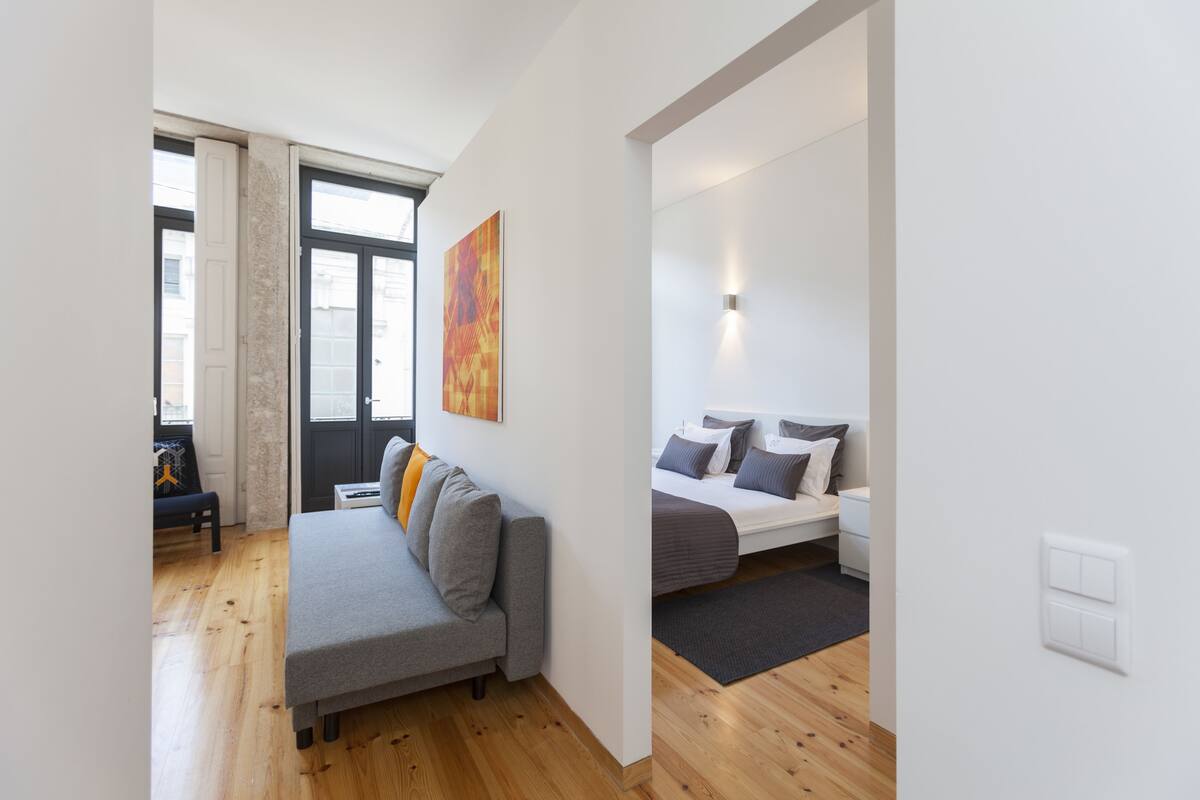Un salon moderne avec un canapé gris, une petite table et un lit avec une literie grise. Il y a une grande fenêtre avec des cadres noirs et un tableau accroché au mur.