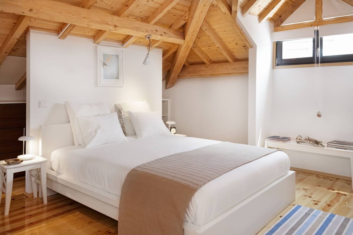 Une chambre avec un grand lit, des poutres apparentes en bois au plafond, une table de chevet avec une lampe, une fenêtre et un petit bureau avec une chaise.