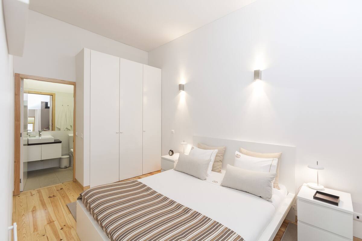Une chambre moderne avec un grand lit, des tables de chevet, une armoire et une salle de bain visible à travers une porte ouverte.