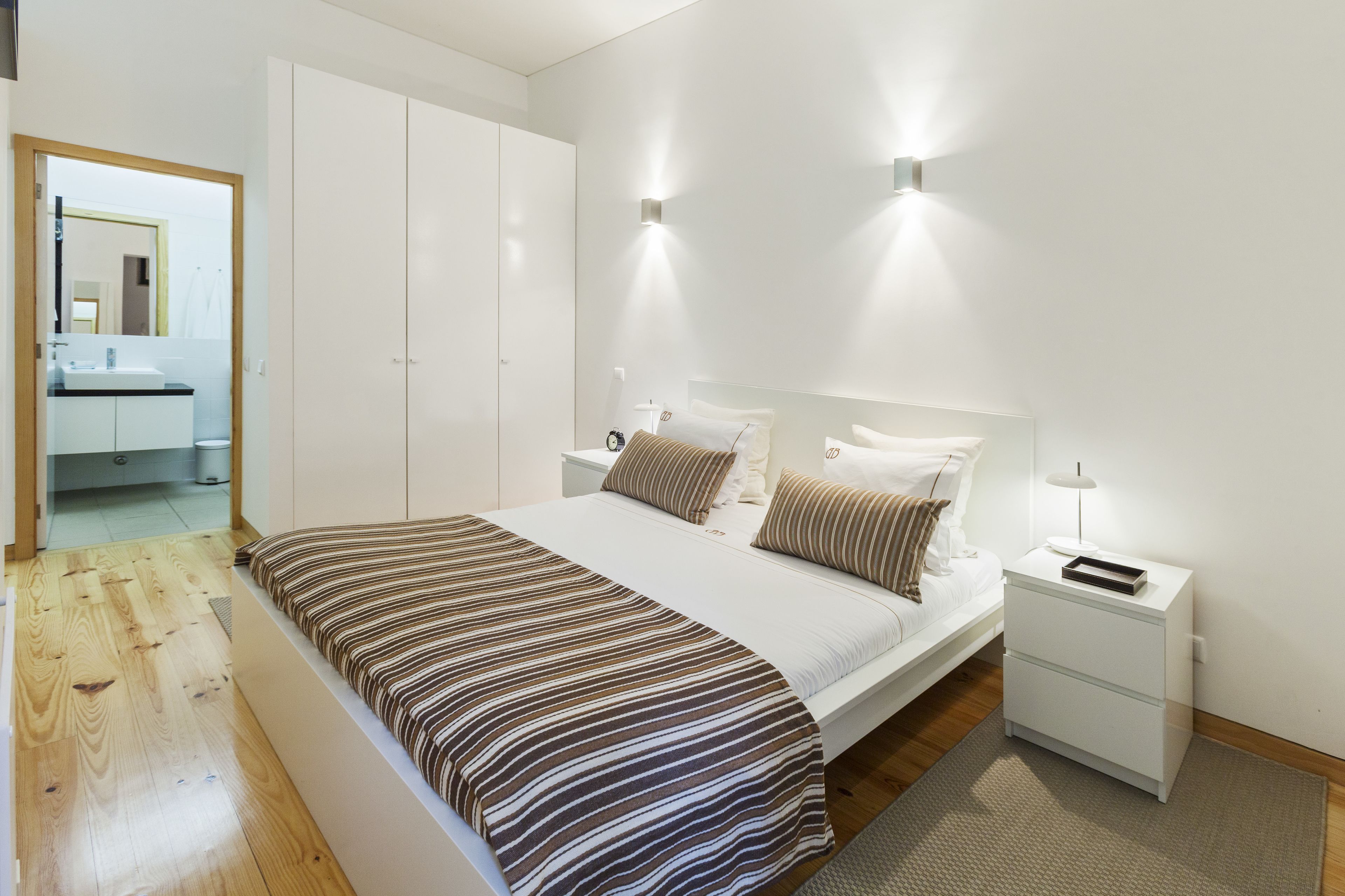 Une chambre à coucher moderne avec un grand lit, une armoire blanche, des tables de chevet et une salle de bain visible à travers une porte ouverte.